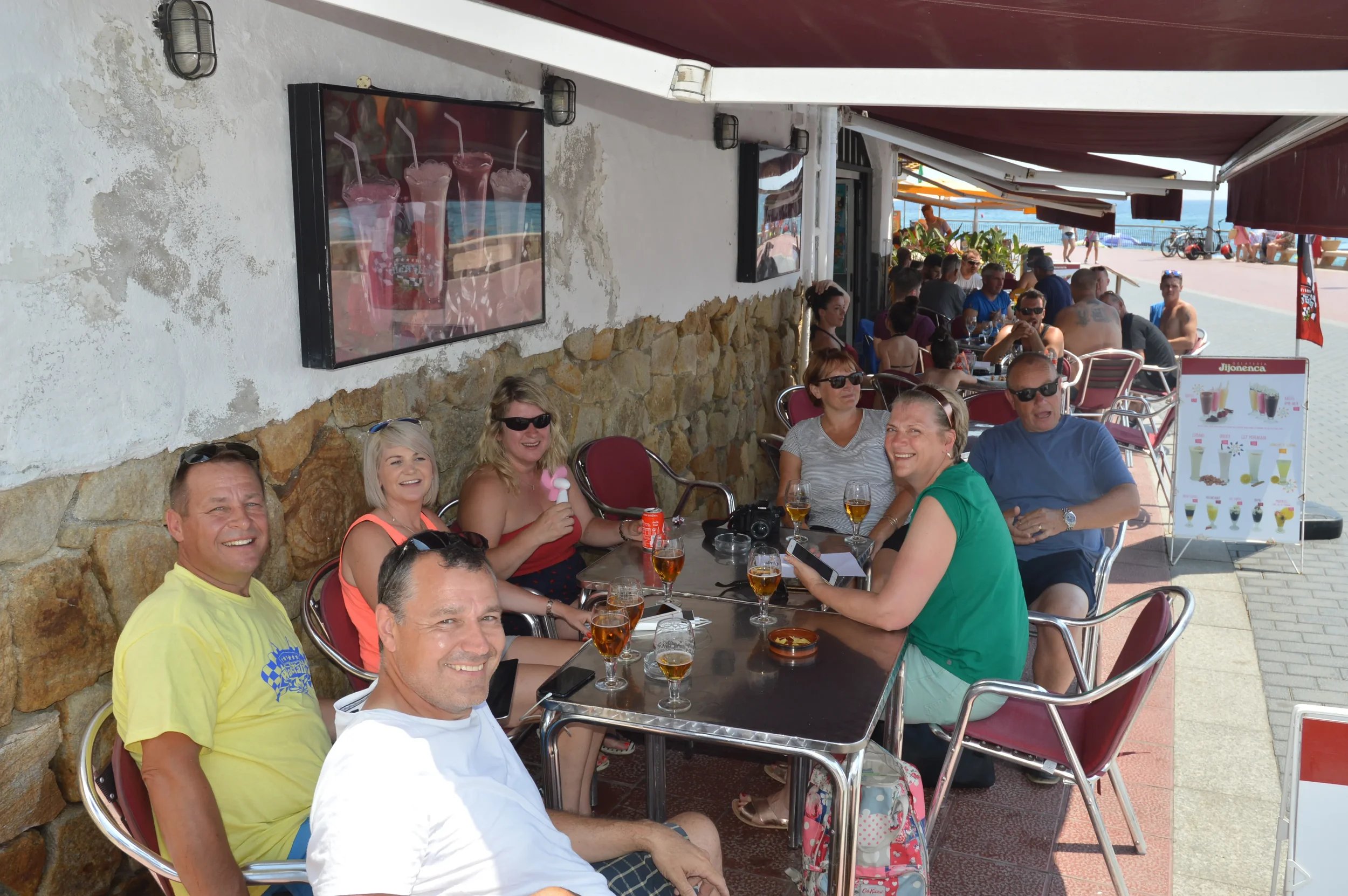 People sitting at an outdoor café with drinks, smiling, enjoying a sunny day near the beach.