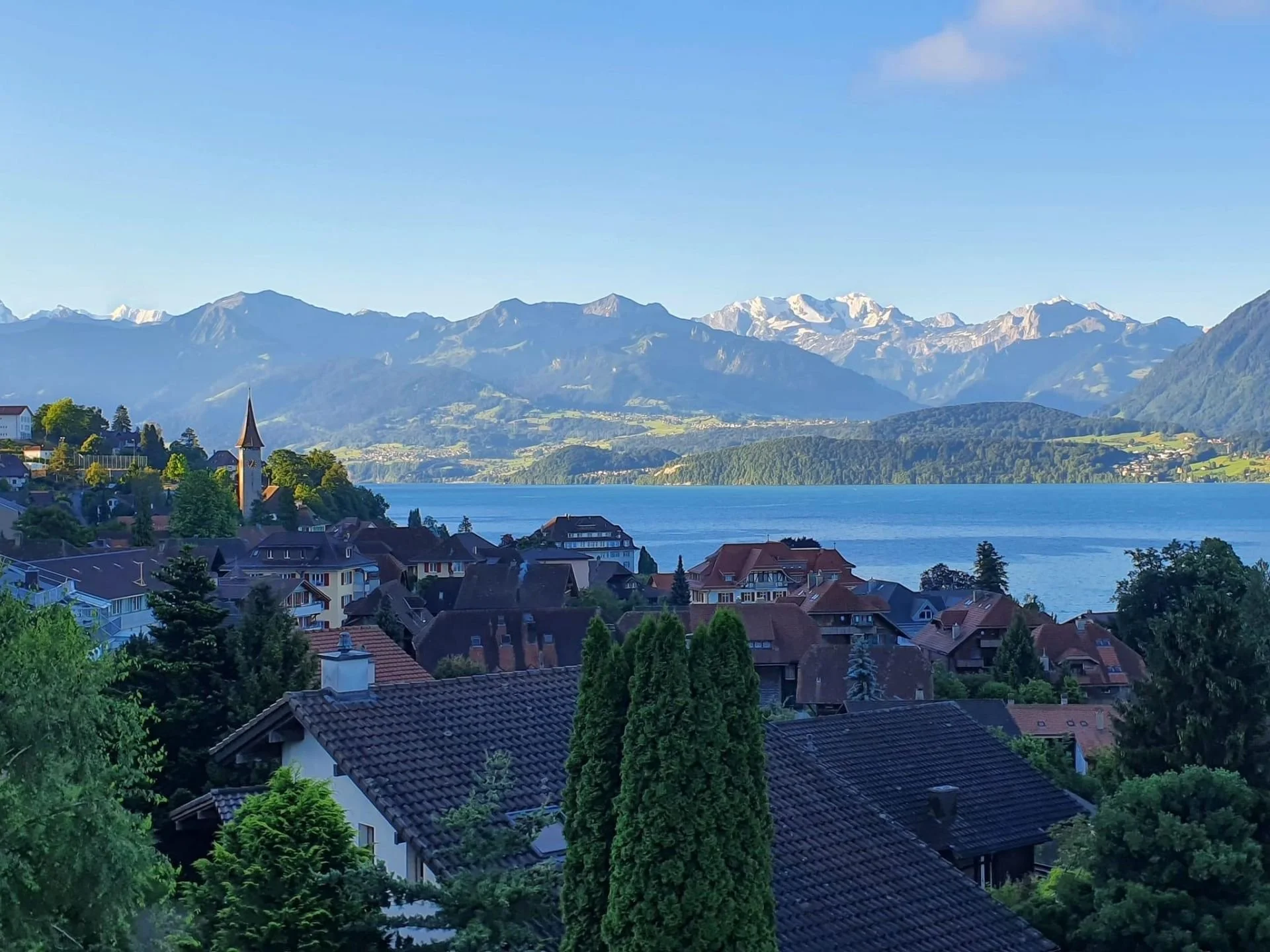 Scenic view of a lakeside village with houses and a church steeple, surrounded by lush green trees, with tall mountains in the background, some snow-capped, under a clear blue sky.