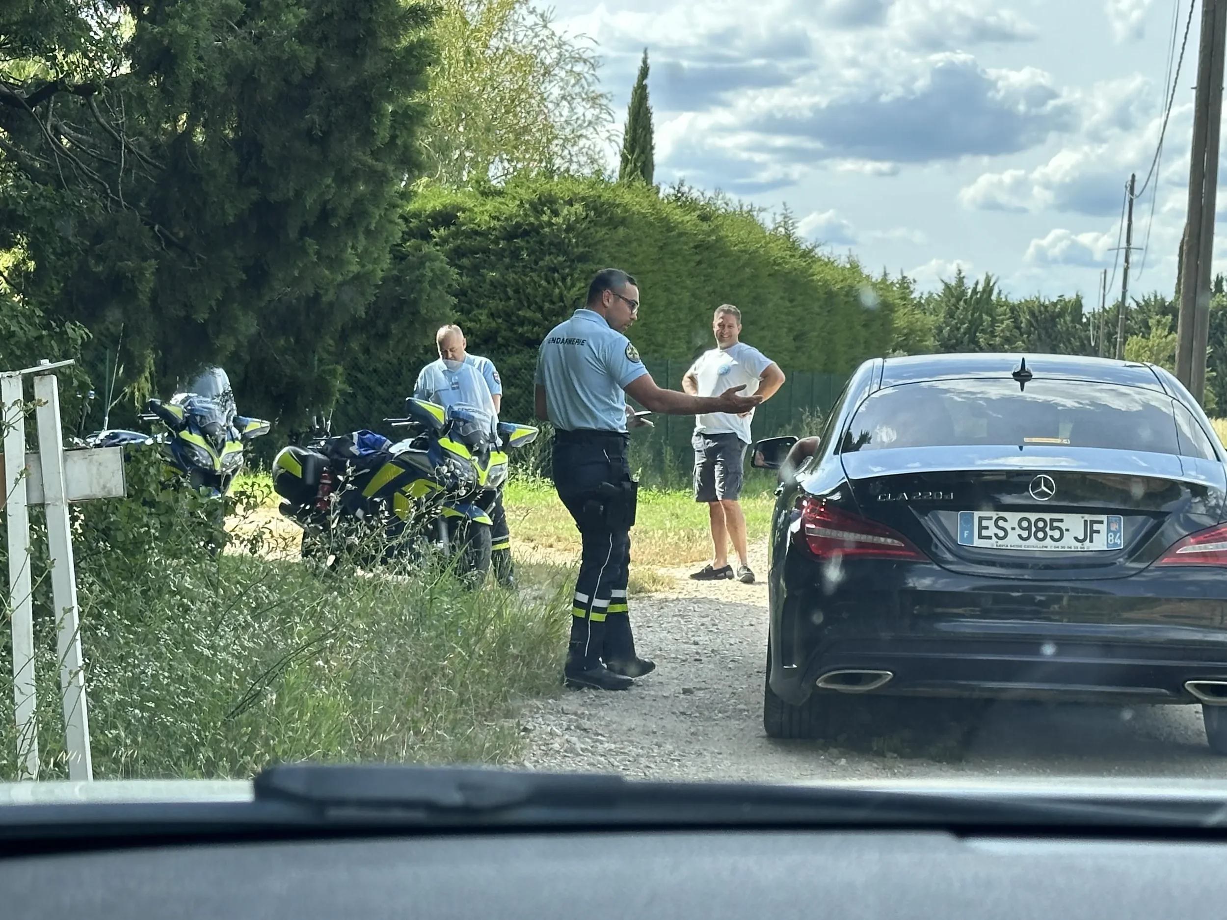 A scene showing two police motorcycles, a police officer speaking to two men near a Mercedes-Benz car, under a partly cloudy sky with trees and power lines in the background.