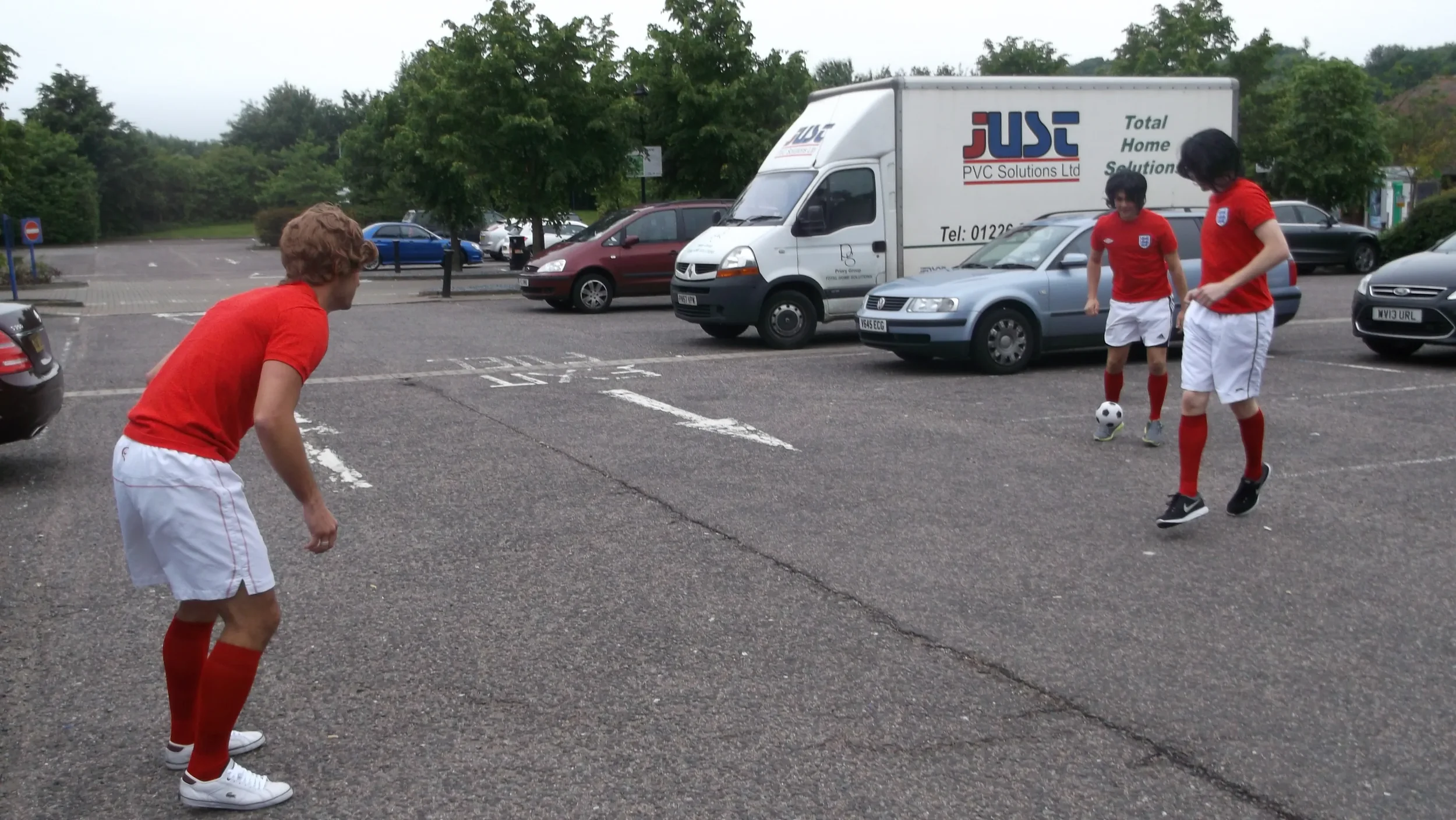 Three young men in red soccer jerseys and white shorts playing with a soccer ball in a parking lot surrounded by parked cars and trees.