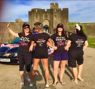 Four women standing in front of a castle, wearing matching black t-shirts with pink and white text, with one woman in purple shorts and another with a flower in her hair, under a partly cloudy sky.