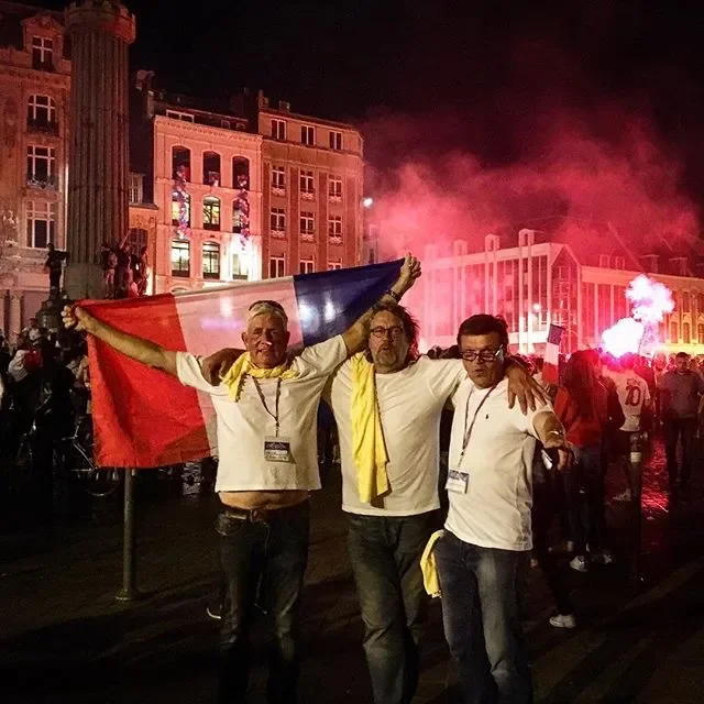 Three men celebrating at a crowded outdoor event at night, holding a French flag with fireworks and fireworks smoke in the background.