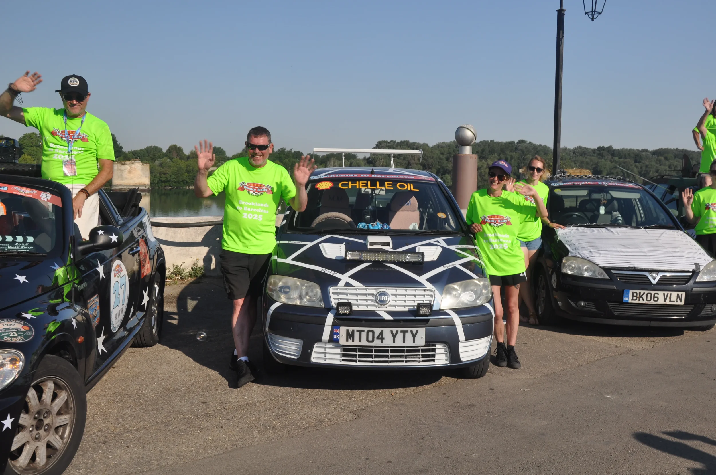 Group of people in bright green shirts standing and waving around decorated cars near a river, with trees and blue sky in the background.