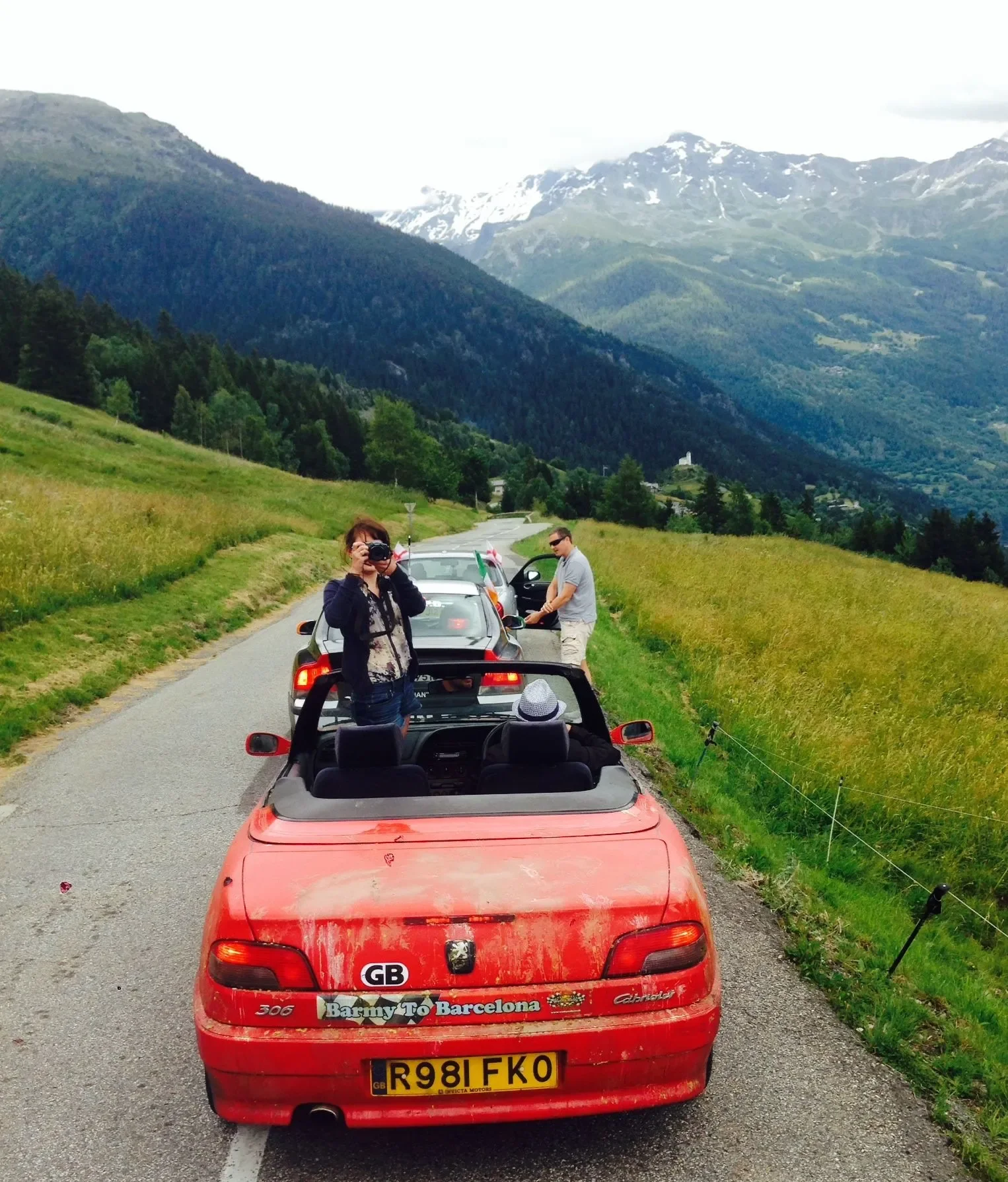 A red convertible car parked on a narrow mountain road with people and cars nearby, surrounded by green fields, trees, and mountains in the background.