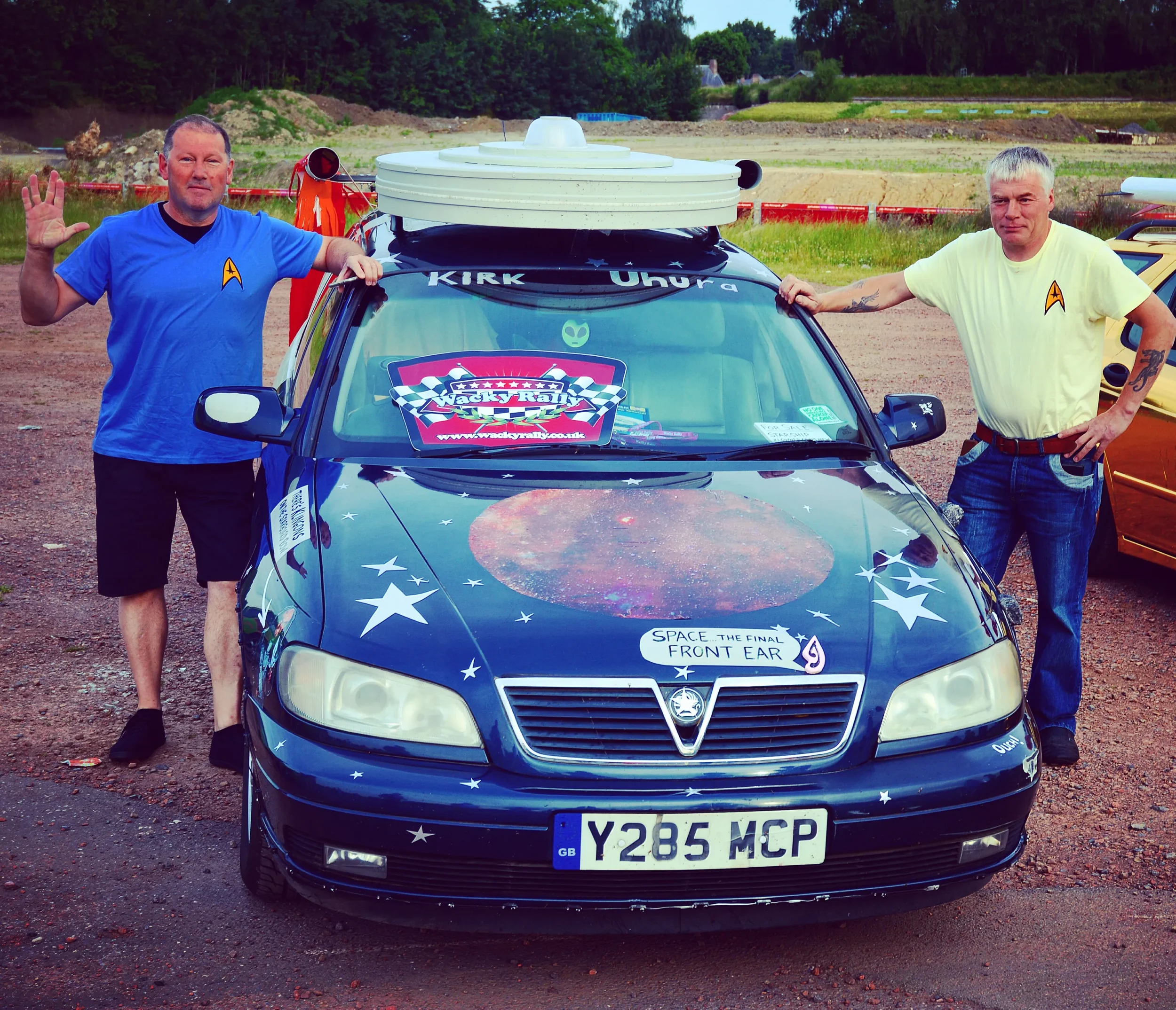 Two men standing next to a UFO-themed decorated car with space-related decals, in an outdoor parking area.