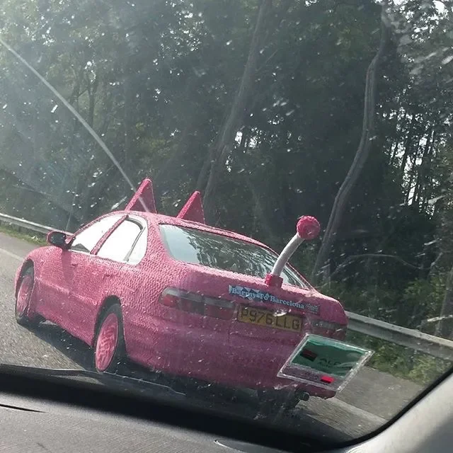 A pink car with a large antenna on the rear, parked on a road surrounded by trees, seen through a dirty windshield.