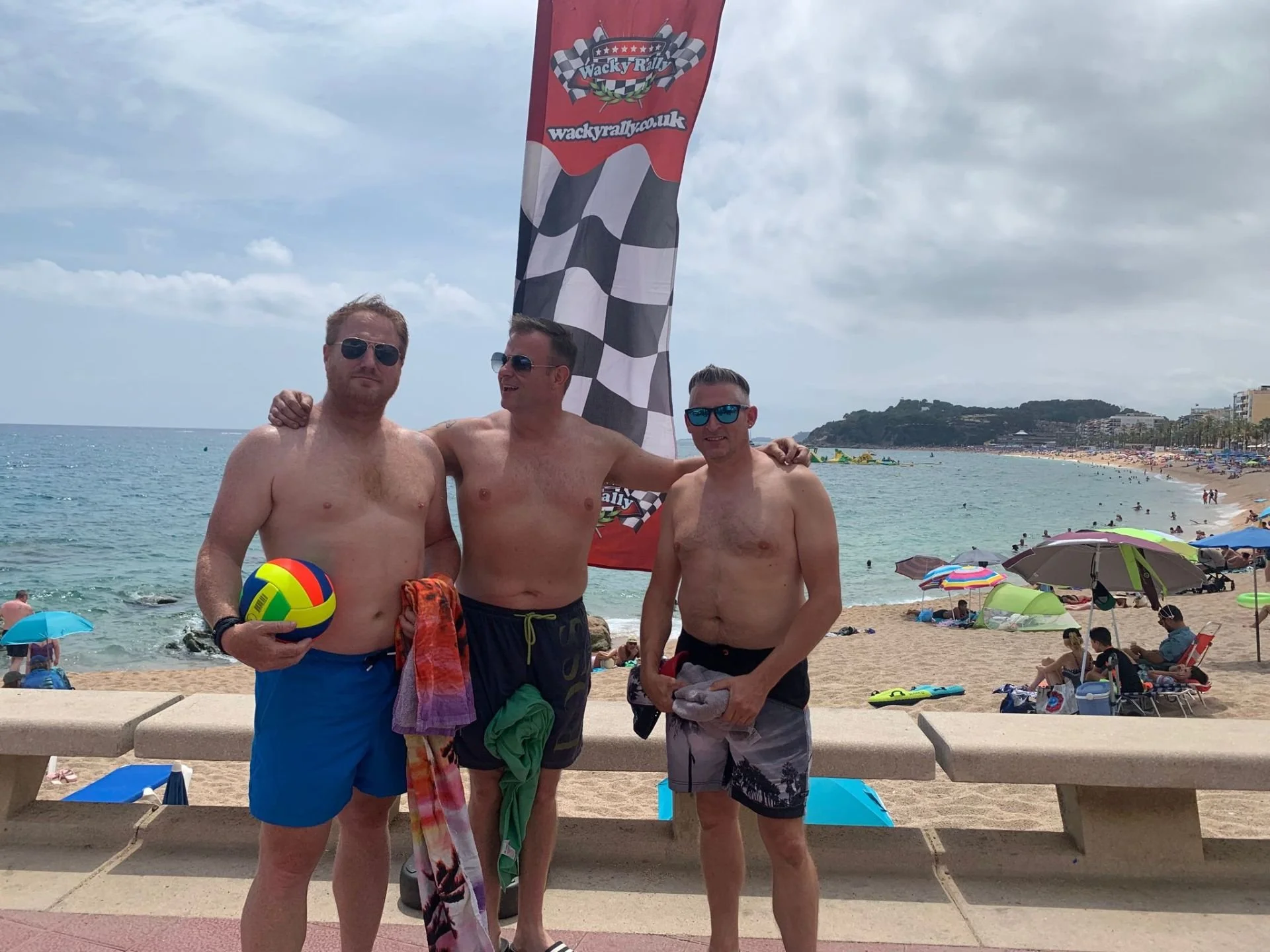 Three shirtless men in swim trunks posing on a beach, with colorful umbrellas, beachgoers, and the ocean in the background. One man holds a volleyball, and there is a checkered flag banner behind them.