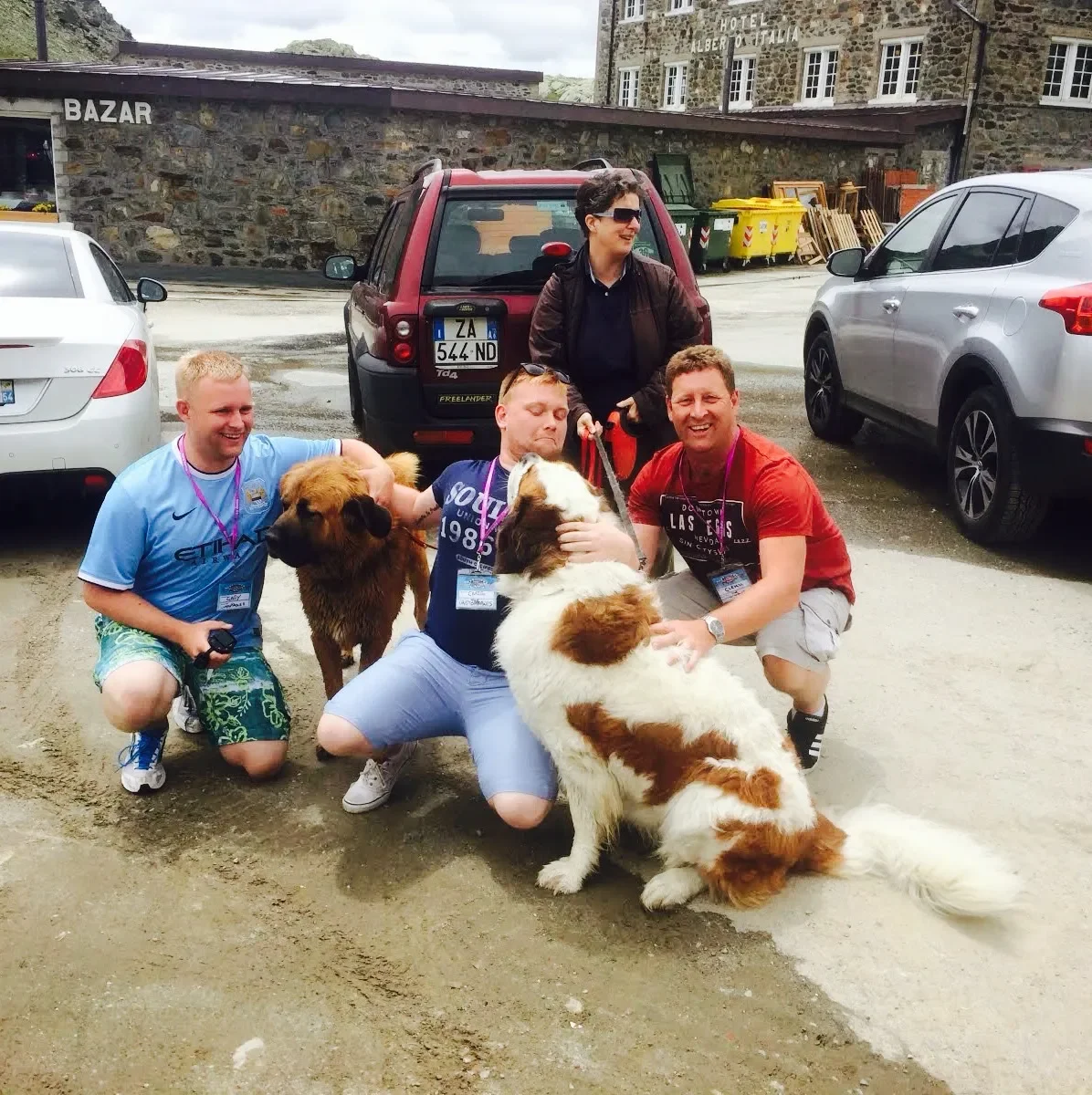 Four people posing with three dogs in a parking lot. A woman is standing behind, and two men are kneeling, holding dogs. One dog is brown with a fluffy coat, and the other is white with brown patches. Several cars are parked, and a building with ston