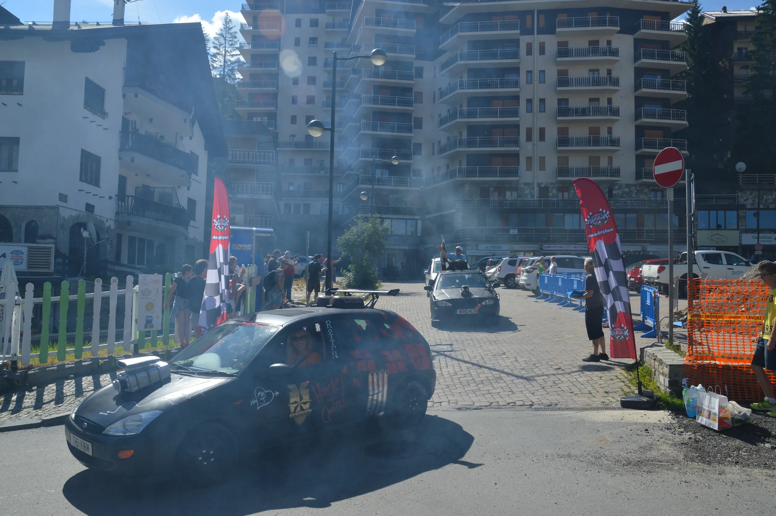 Car race event with vintage cars, people gathered along the street, and hotel buildings in the background.