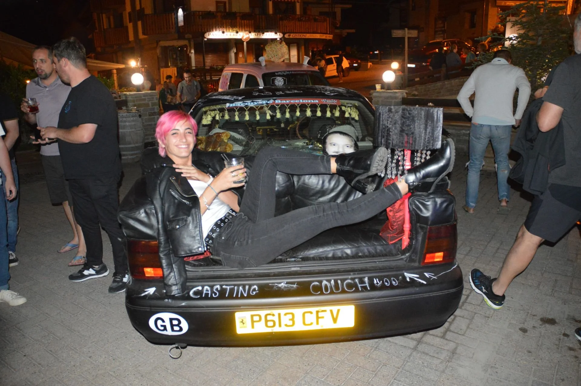 Person with pink hair lying in the trunk of a car, smiling and holding a drink, surrounded by people at an outdoor event at night.