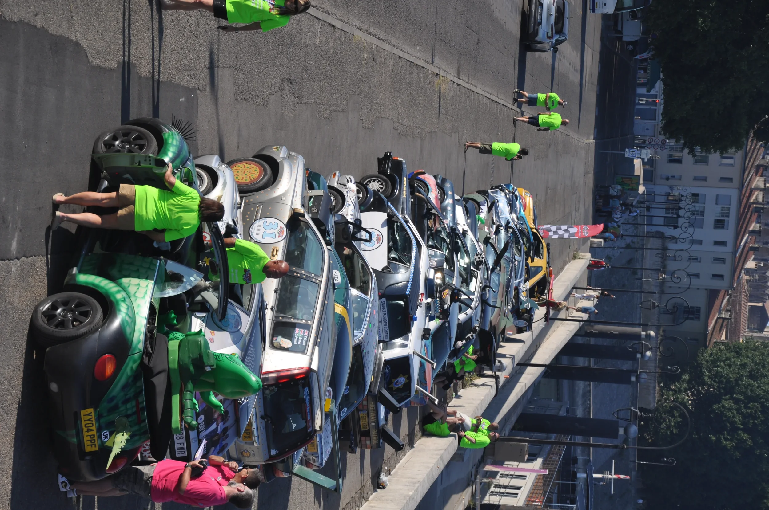 A row of classic cars and vintage vehicles parked along a city street with several people in green t-shirts walking and inspecting the cars, and a few pedestrians on the sidewalk.