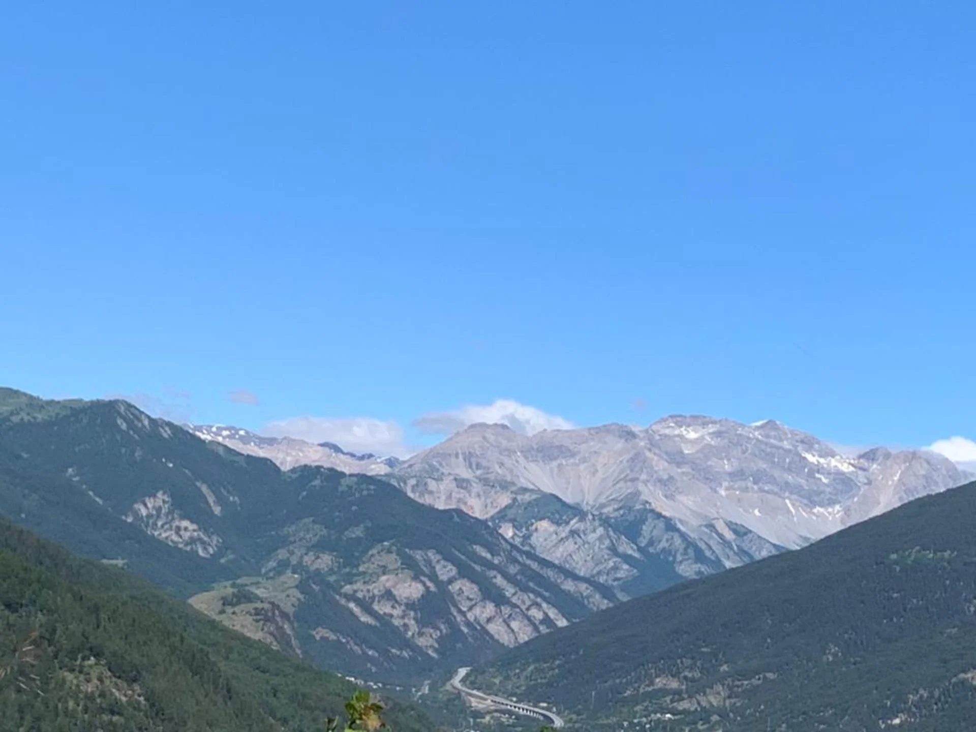 Scenic view of mountains with green forests and snow patches, under a blue sky.