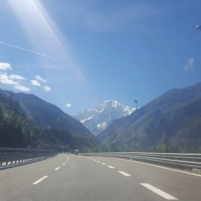 A clear highway road with mountains, including a snow-capped peak, in the background under a bright blue sky with some clouds and sunlight.