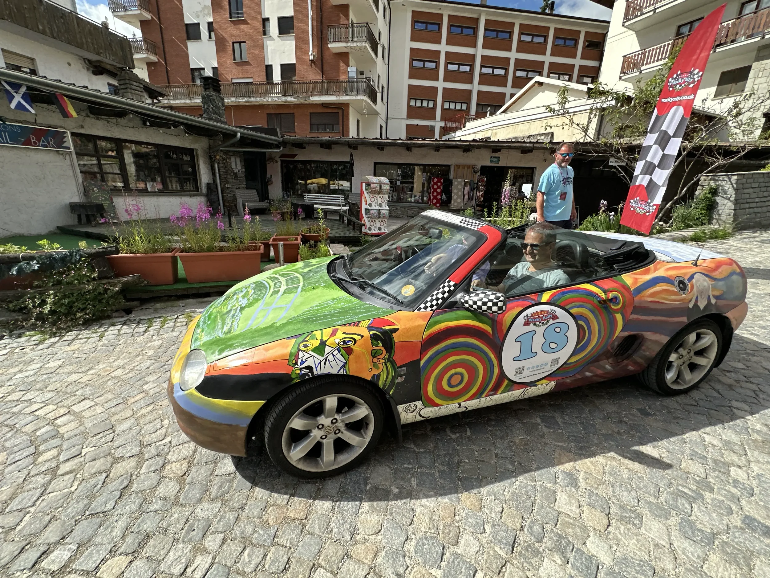 A colorful sports car with an abstract painted exterior, featuring swirls and faces, parked on cobblestone street. Two men are inside the car, with one wearing sunglasses. A third man stands beside the car; a tall flag with checkered pattern and logo