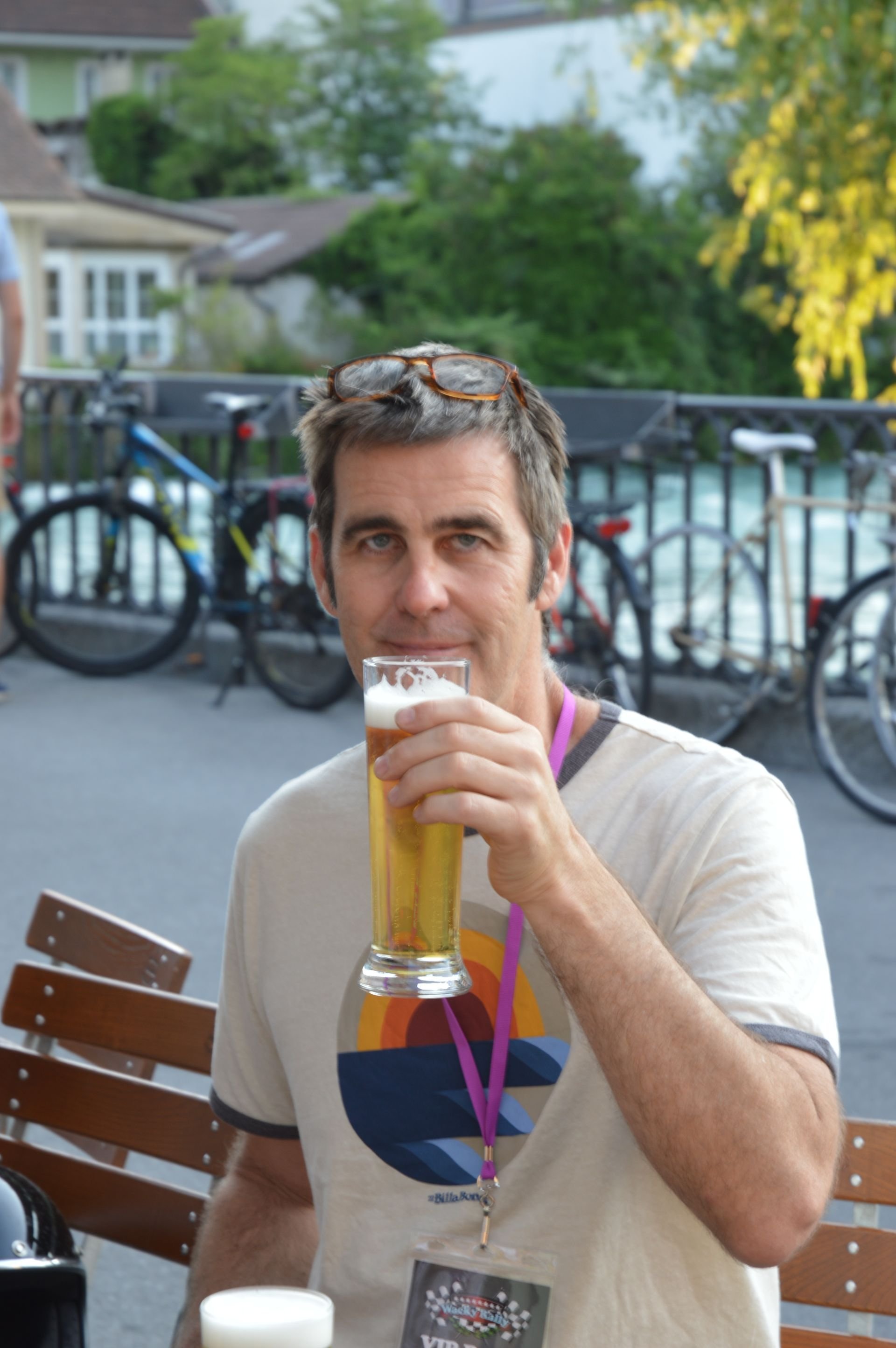 Man outdoors holding a glass of beer, wearing glasses on his head and a lanyard with an ID badge around his neck, with bicycles and green trees in the background.