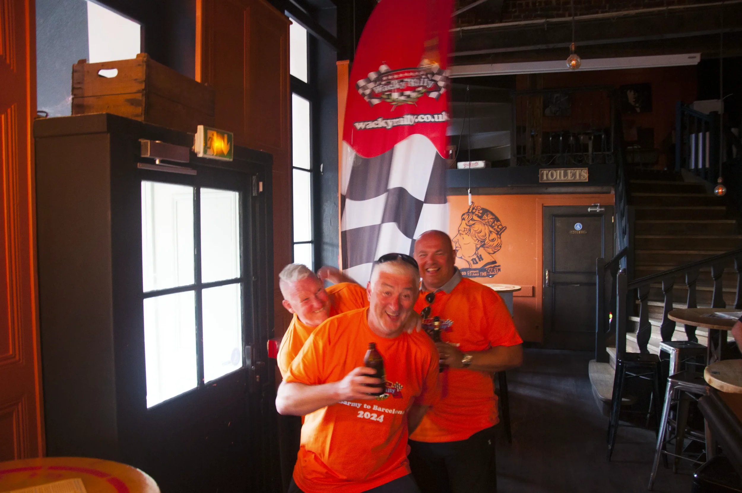 Three smiling men in orange T-shirts celebrating near a checkered flag banner inside a bar or restaurant. One man is holding a bottle. The background shows stairs, a 'TOILETS' sign, and a wall mural of a woman with a sign beneath it.