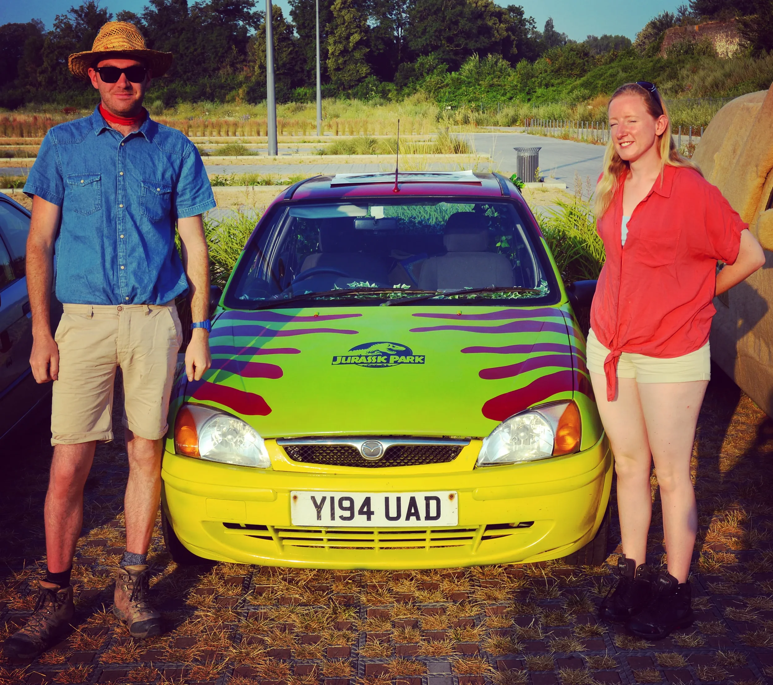 Two people standing next to a colorful 'Jurassic Park' themed Mazda car in an outdoor parking area with greenery in the background.