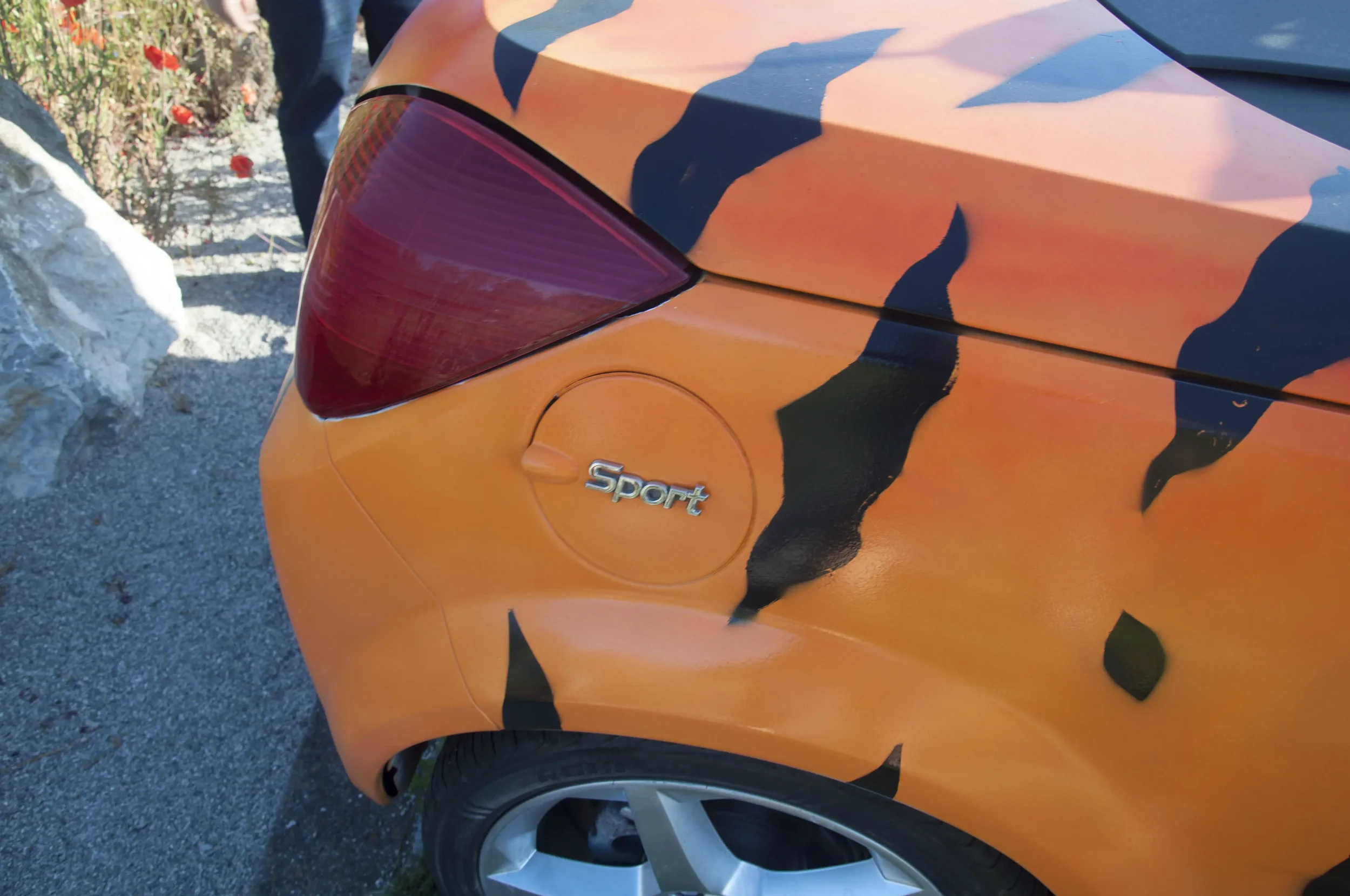 Rear side of a sports car painted in orange with black stripe camouflage pattern, showing the taillight, the 'Sport' badge, and part of the rear wheel.