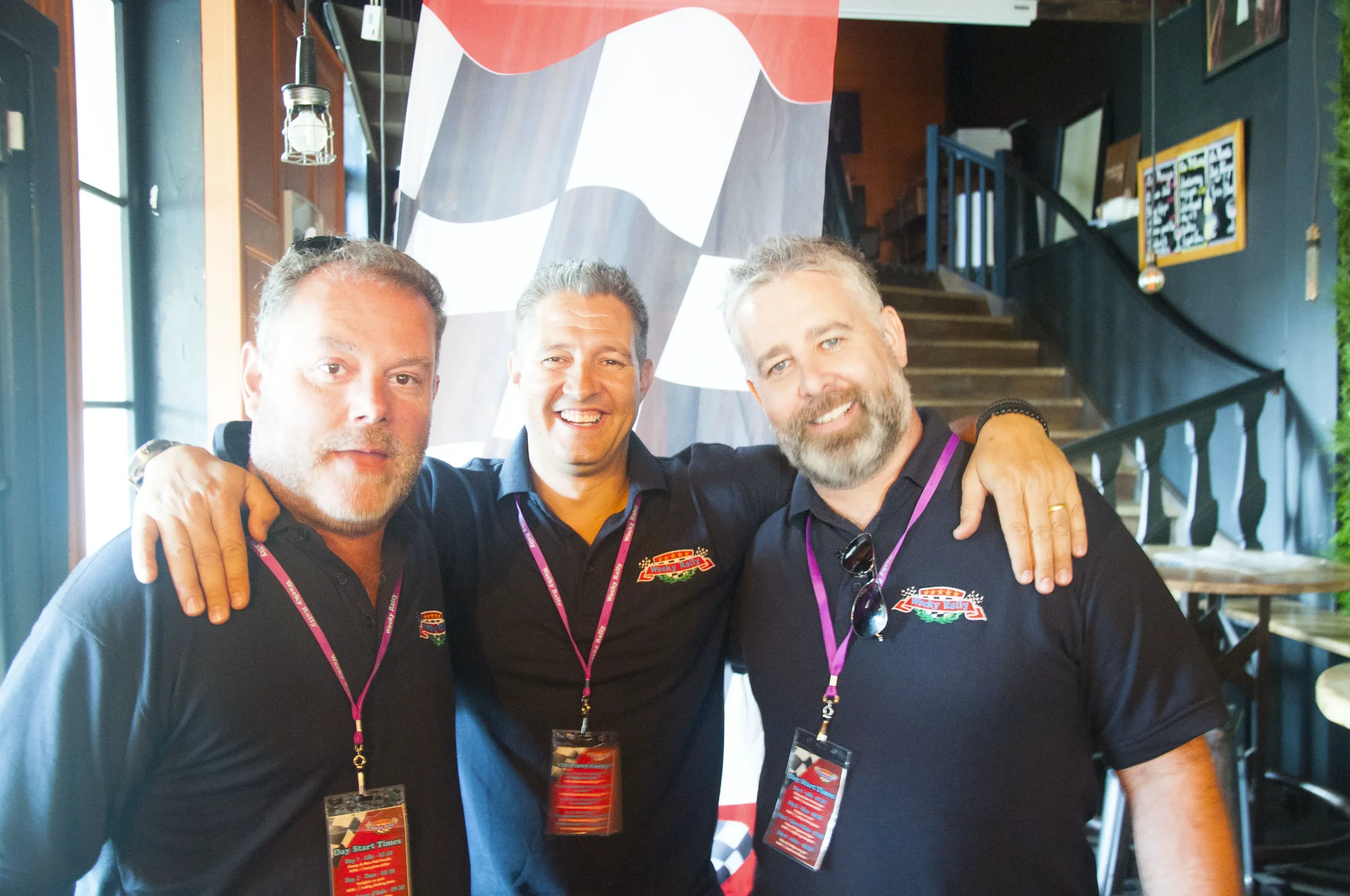 Three smiling men with their arms around each other's shoulders, wearing black shirts with a logo, standing indoors with a staircase in the background, and a large checkered flag banner behind them.