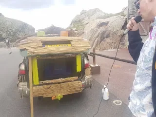 Person holding a microphone next to a vehicle with a makeshift wooden structure on the back, set against a rocky outdoor landscape.
