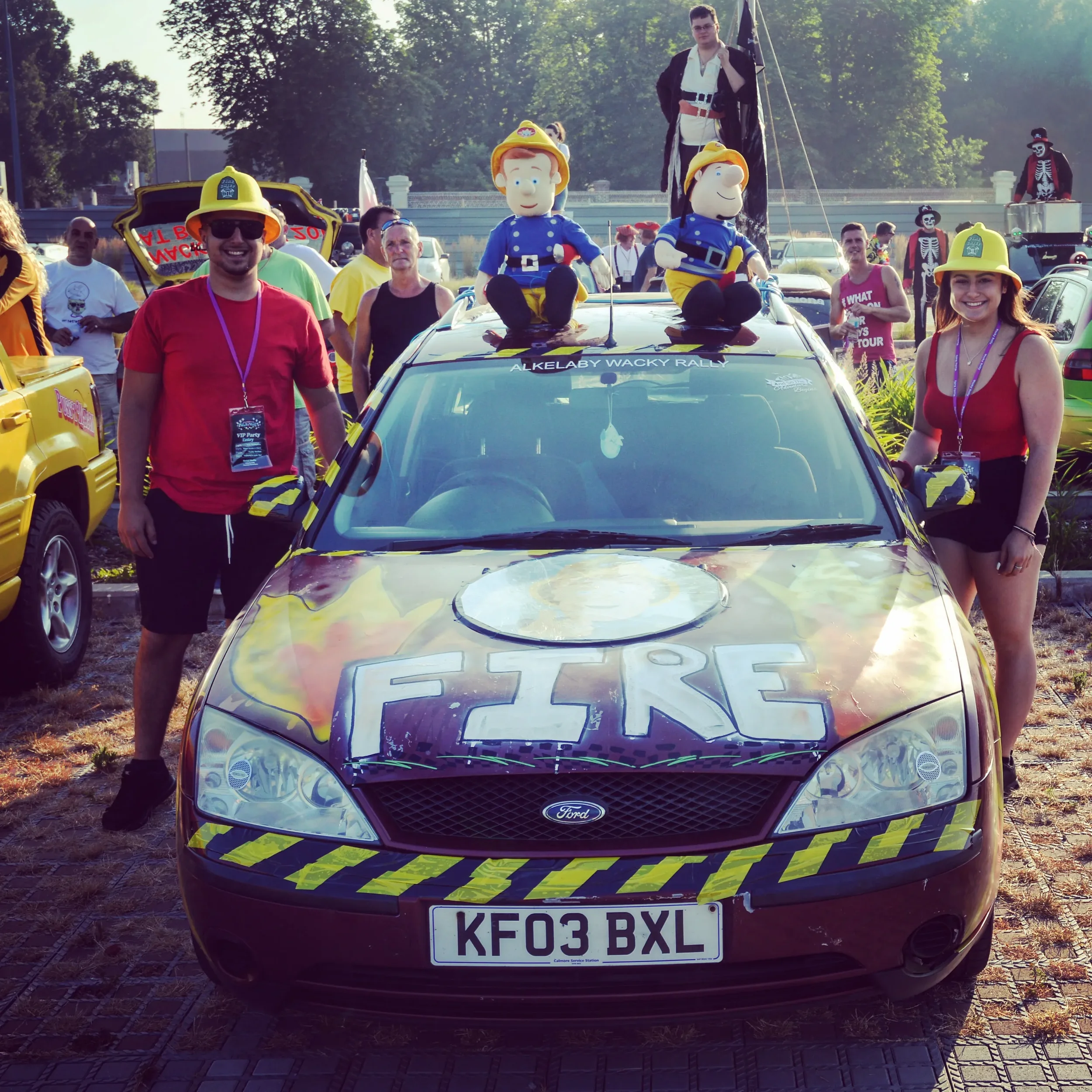 A group of people dressed as firefighters surrounding a decorated Ford car with firefighter helmets on the hood, two plush toy firefighters on top of the car, and other costumed individuals in the background during a parade or event. The front of the