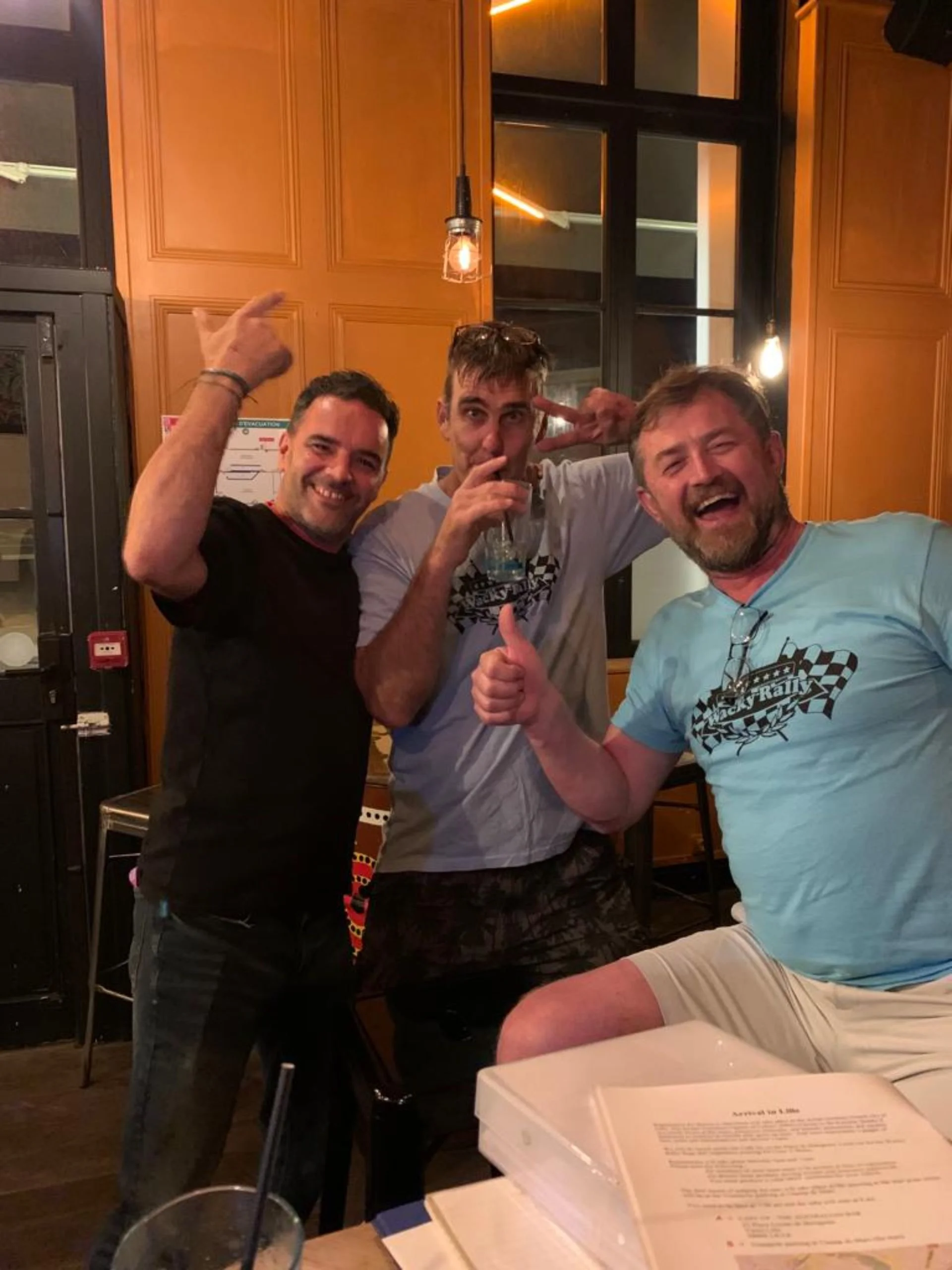 Three men happy and celebrating indoors, with one holding a drink and showing peace sign, another giving a thumbs up, all smiling.