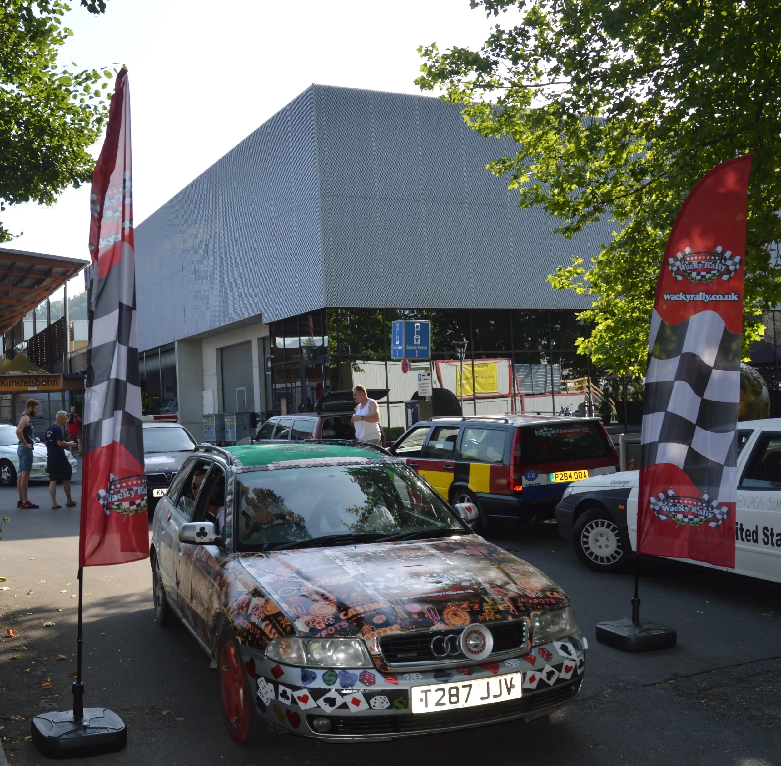 A decorated car with playing card motifs parked outdoors under trees, flanked by two red flags with checkered pattern and a 
