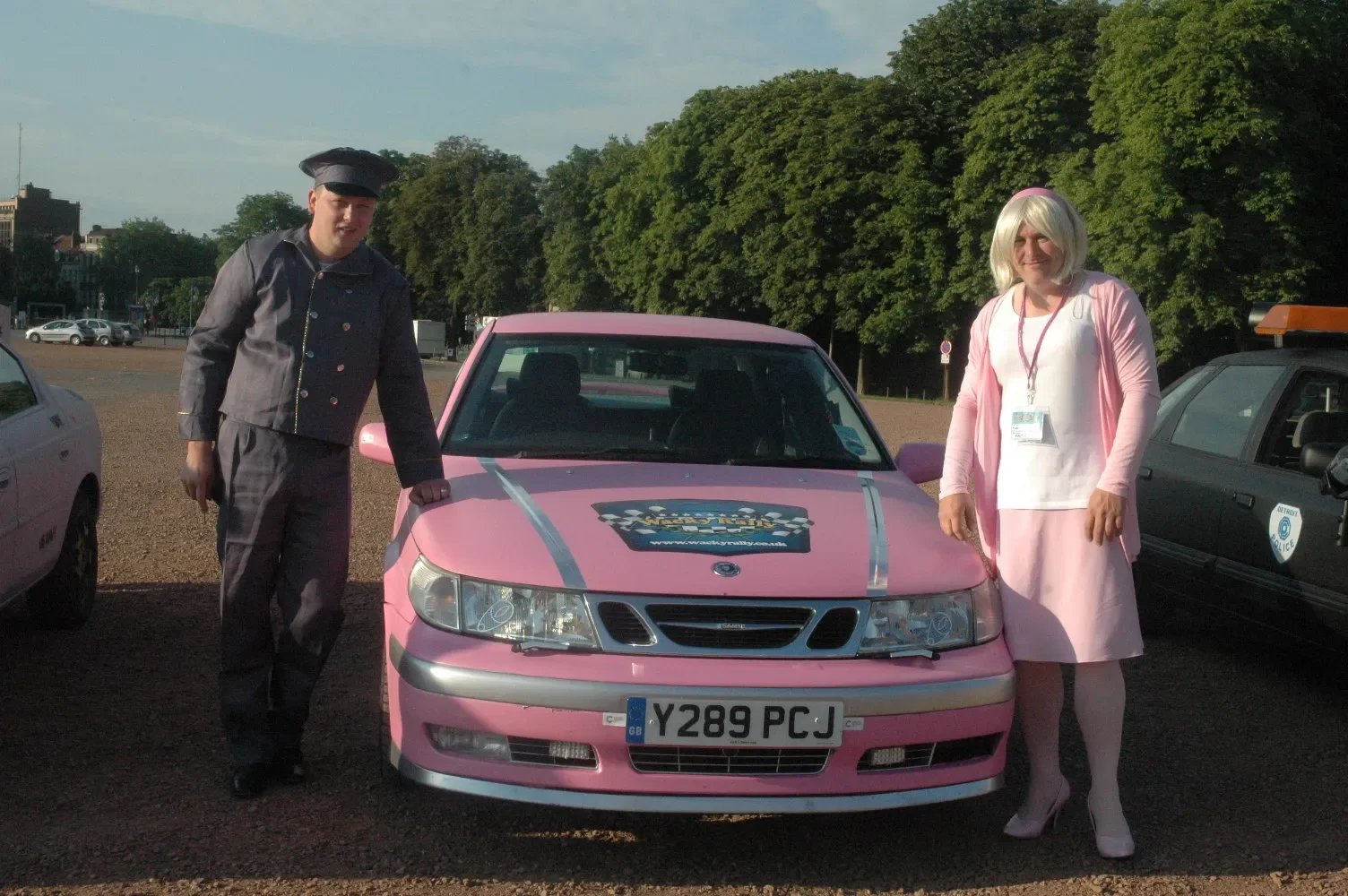 Two people dressed in costumes stand next to a pink car with a checkered flag decal on the hood. The person on the left is in a gray police badge and tie, and the person on the right is in a pink dress, white sash, and a wig with blonde hair.