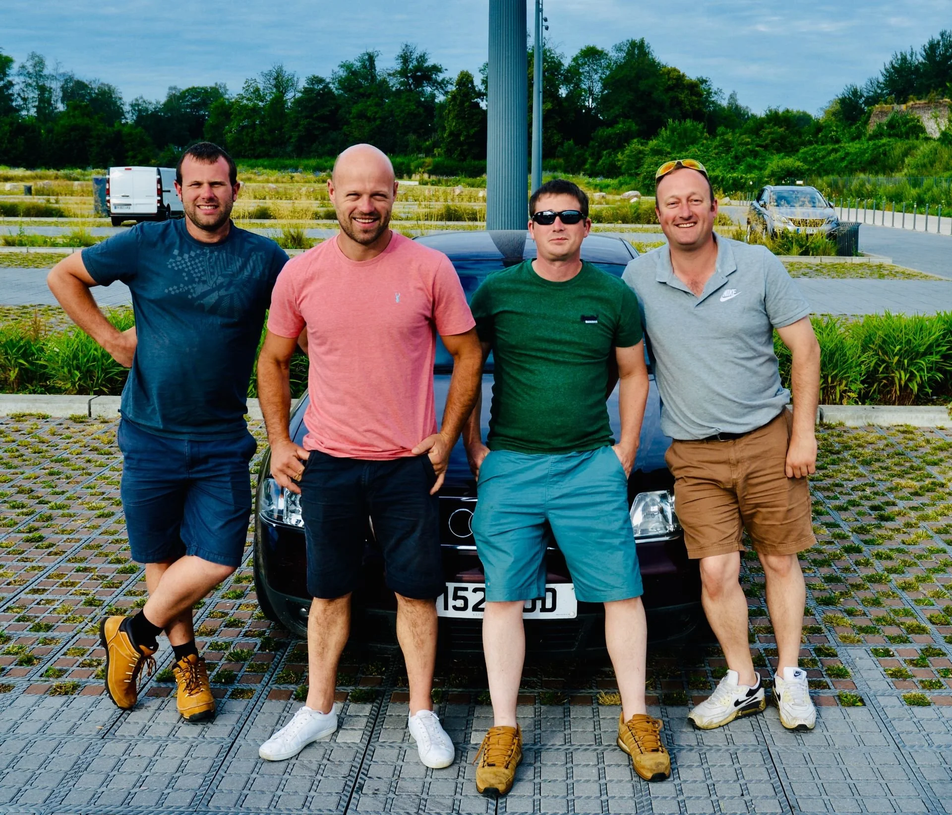 Four men standing in front of a black car in a parking lot with greenery and trees in the background.