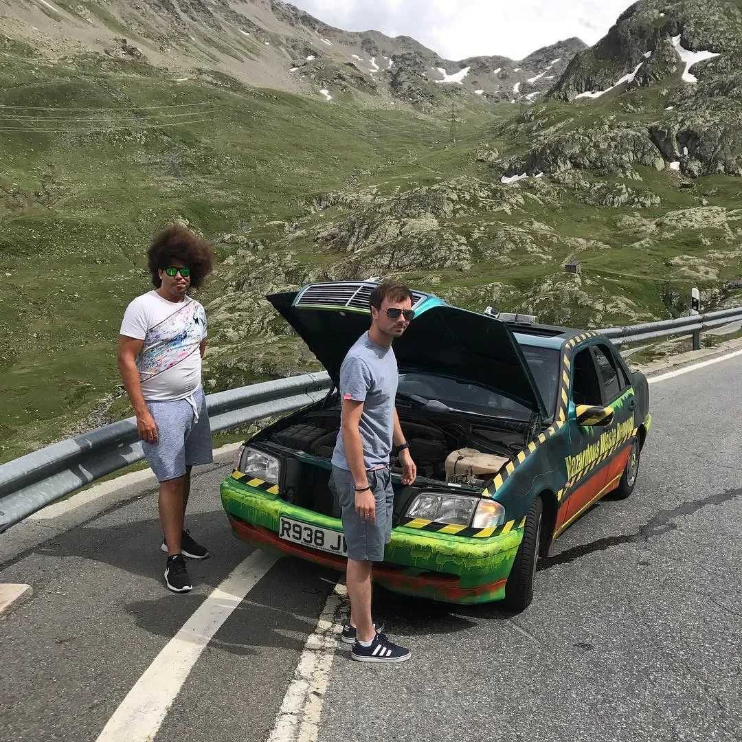 Two young men standing by a car with its hood open on a mountain road. The car has a colorful, graffiti-style paint job. The scene is set in a mountainous landscape with green hills and rocky peaks.