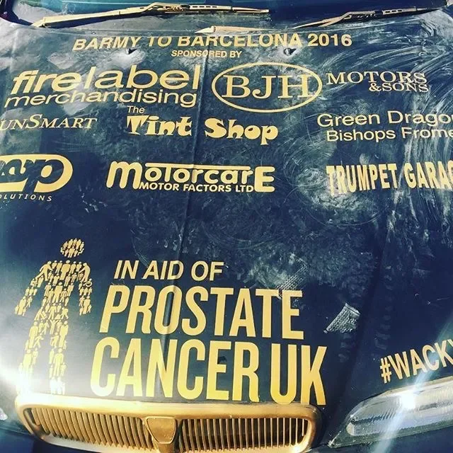 Close-up of a car hood with various sponsor logos and messages supporting a charity event for prostate cancer UK, including a large yellow message and a hashtag.