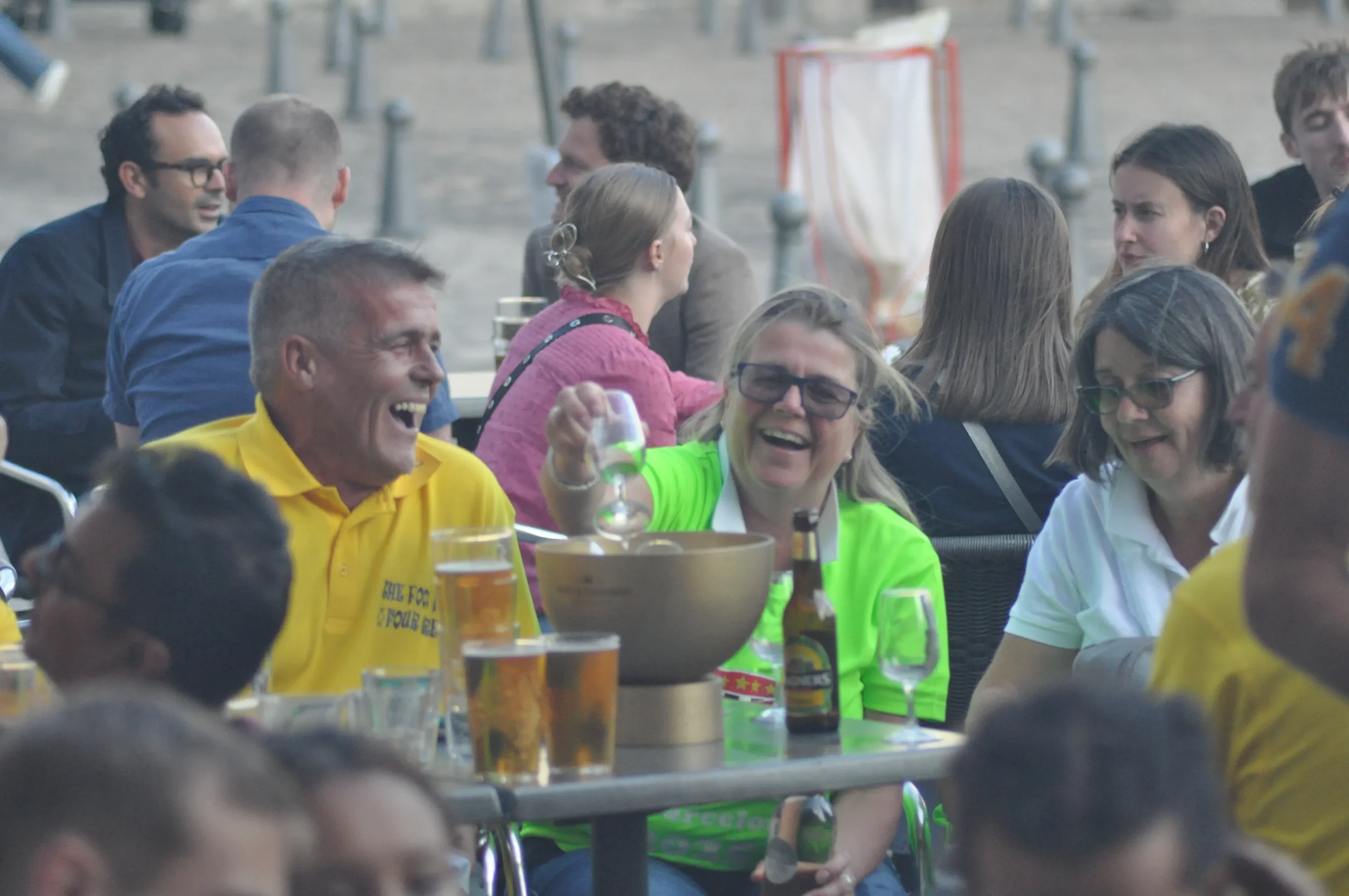 People sitting at outdoor tables, laughing and enjoying drinks at a social gathering, with some individuals wearing bright clothing.