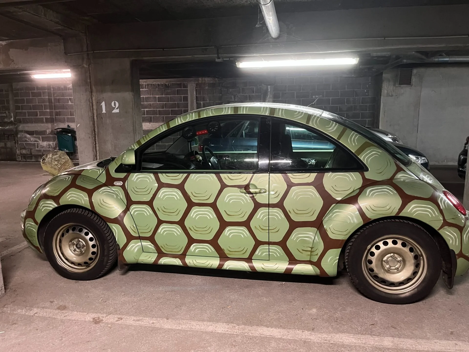 A Volkswagen Beetle car with a honeycomb pattern wrap in green and brown in a parking garage.