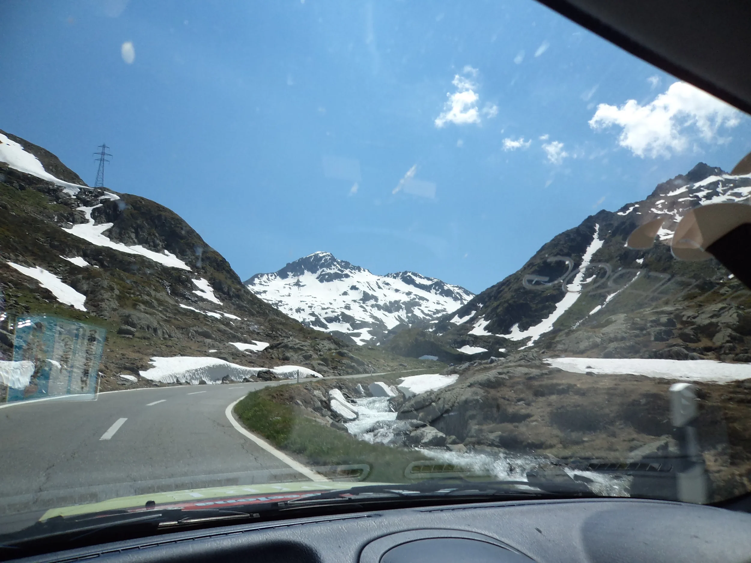 A mountain road with snow-capped peaks on either side, taken from inside a vehicle with a clear view of the windshield and dashboard, showing blue sky with a few clouds.