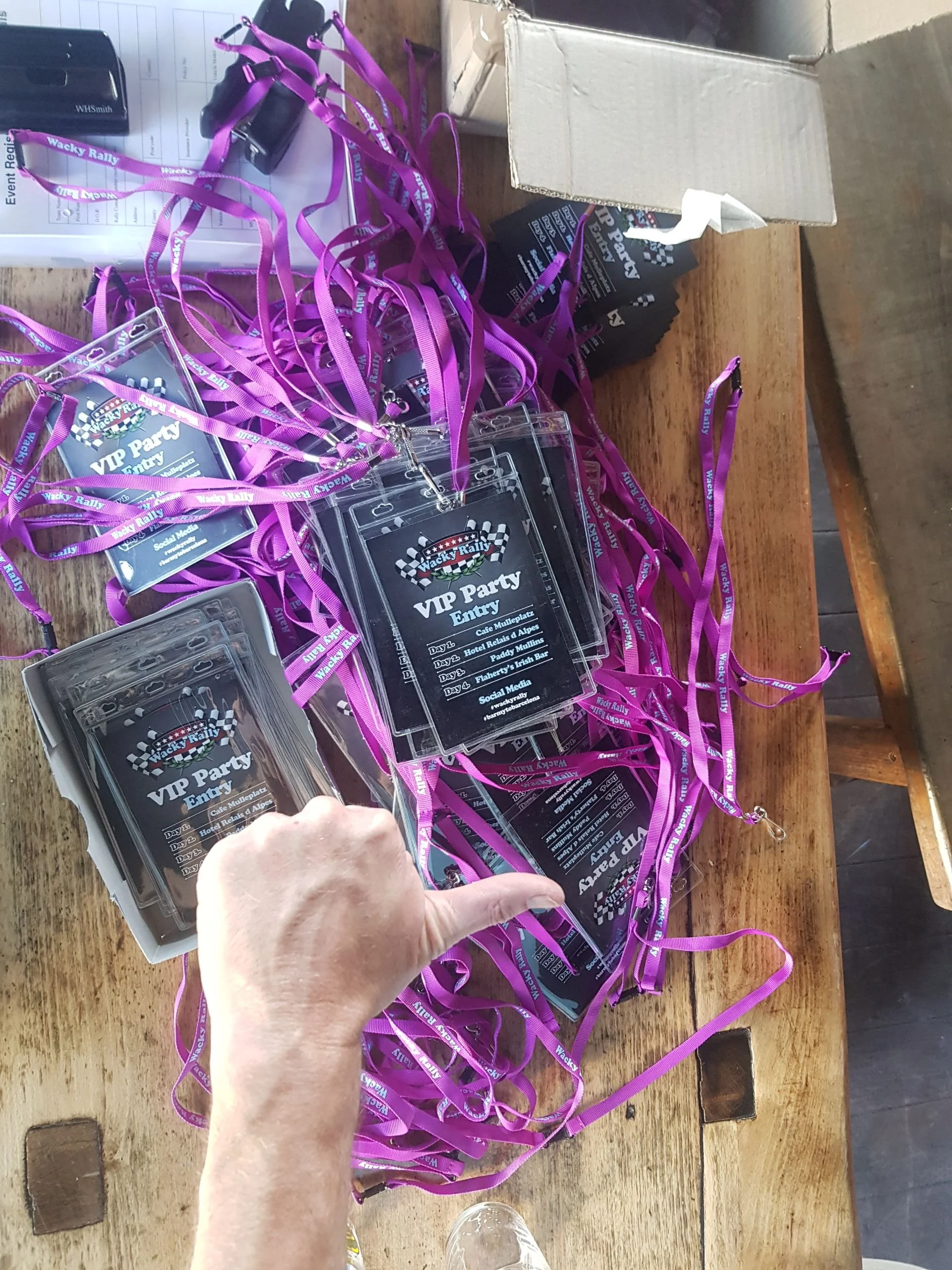 A person's hand pointing at a pile of Wacky Rally VIP party passes with purple lanyards on a wooden table.