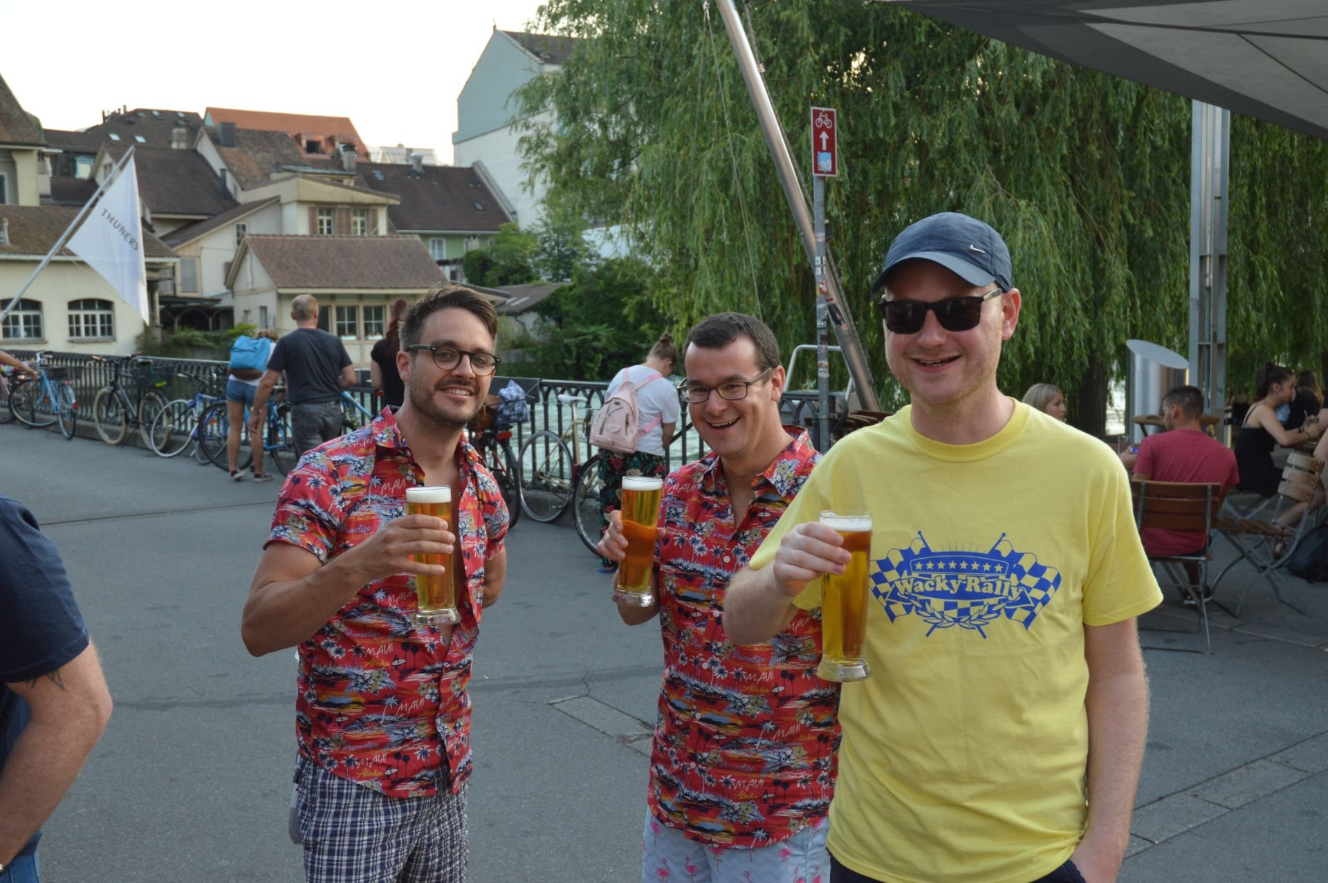 Three men smiling and holding glasses of beer outdoors near a river with bicycles and other people in the background.