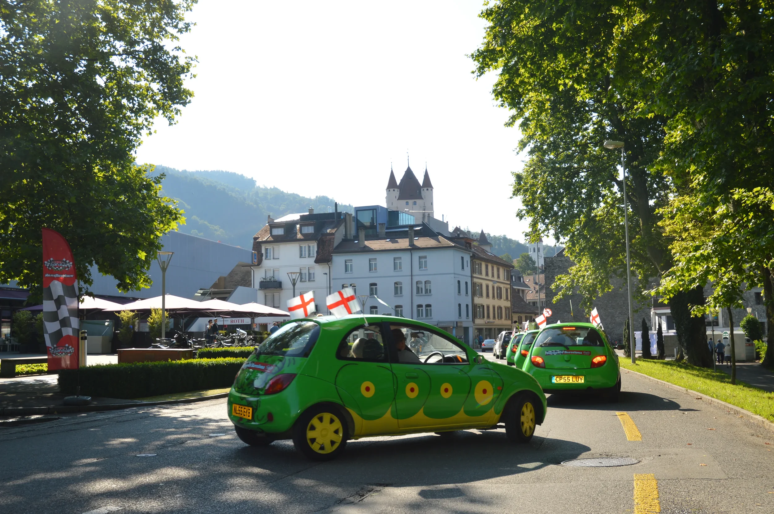 Green car with yellow wheels and race car decals on a street lined with trees, with more similar cars in the background, and a castle on a hill in the distance.