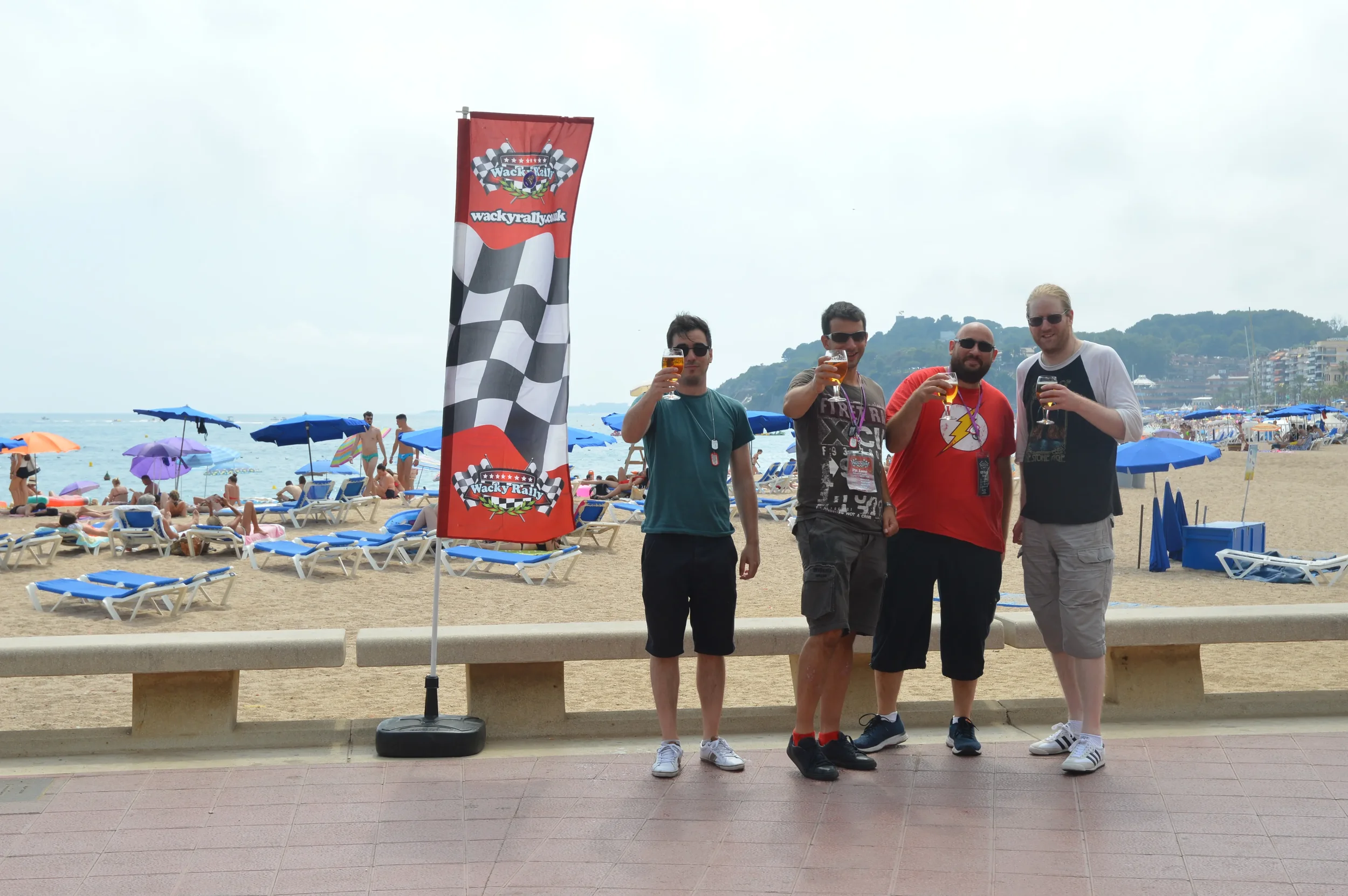 Four men standing on a promenade at the beach, holding beverages, with a flag that has a checkered race flag pattern and the logo for Wackyrally, in front of a crowded beach with umbrellas and people, and a hilly coastline in the background.