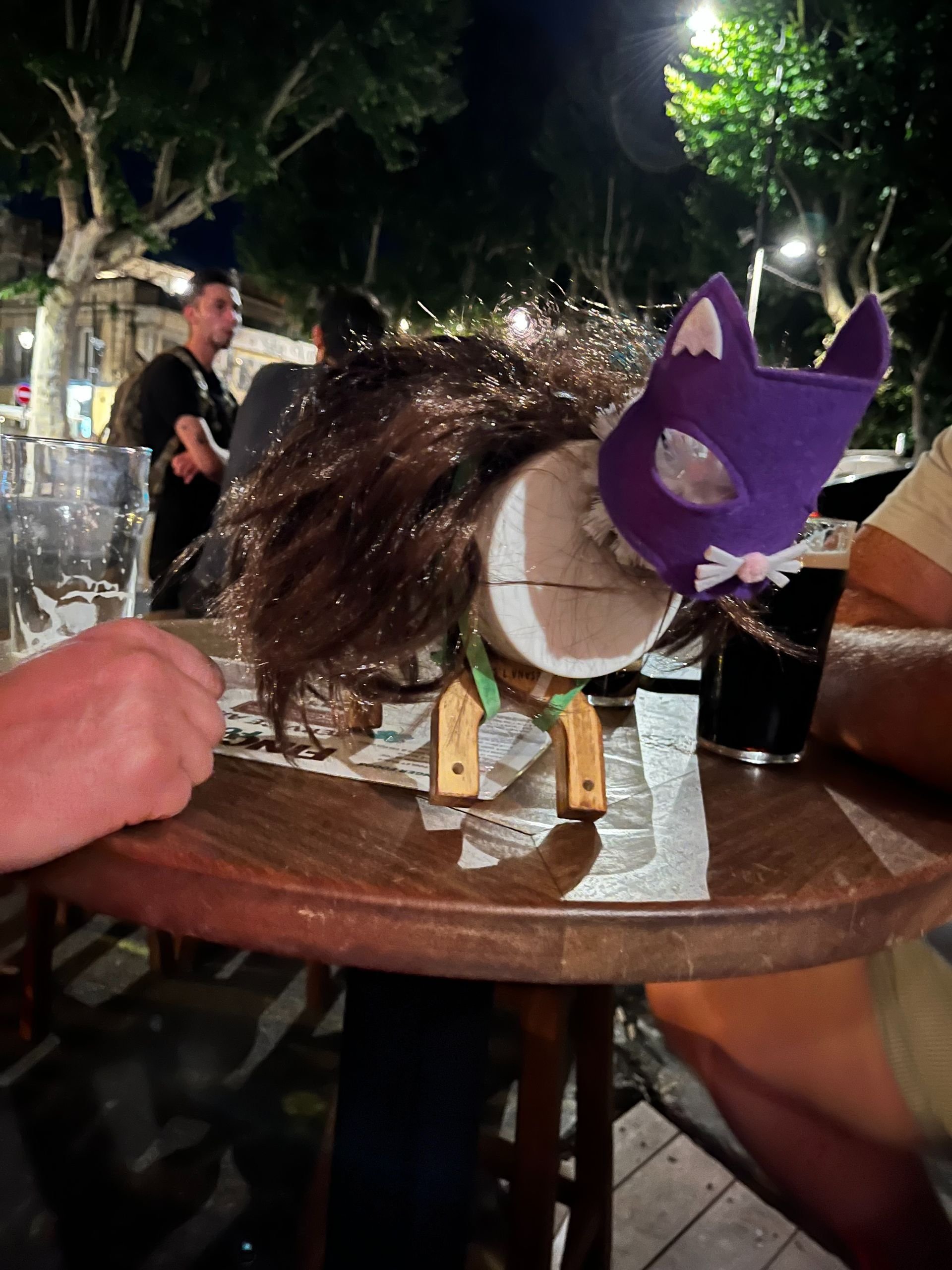 A table with a decorative cat figurine wearing a purple mask and a wig, surrounded by drinks at an outdoor nighttime setting. There are people in the background.