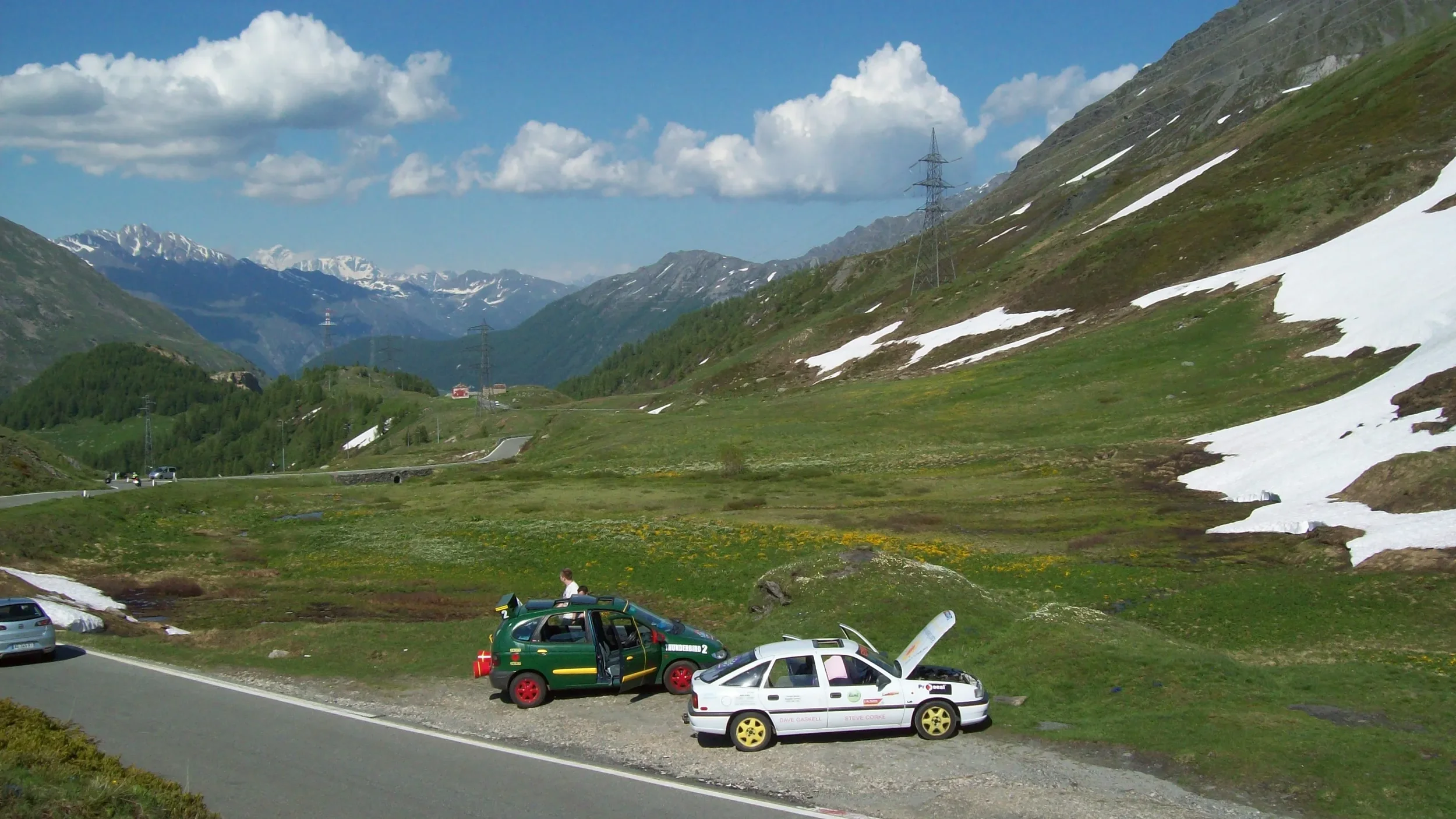 Two rally cars parked on a grass roadside in a mountainous landscape with patches of snow, green hills, and distant snow-capped mountains under a partly cloudy sky.