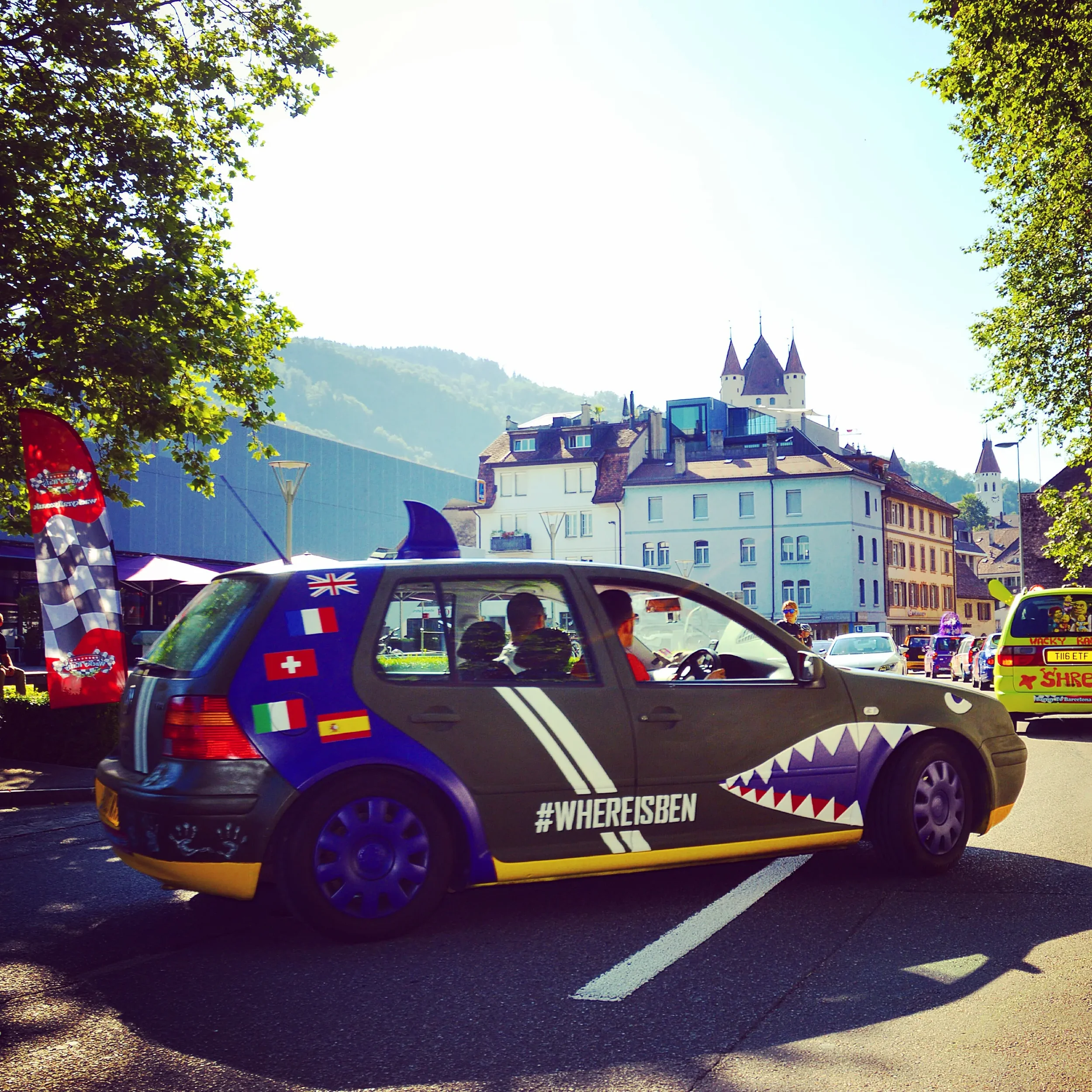 A decorated car with shark teeth graphics and country flags parked on a street in a European town with historic buildings and mountains in the background.