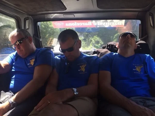 Three men wearing blue shirts with a yellow eagle emblem, sleeping in the back of a vehicle with sunlight outside.