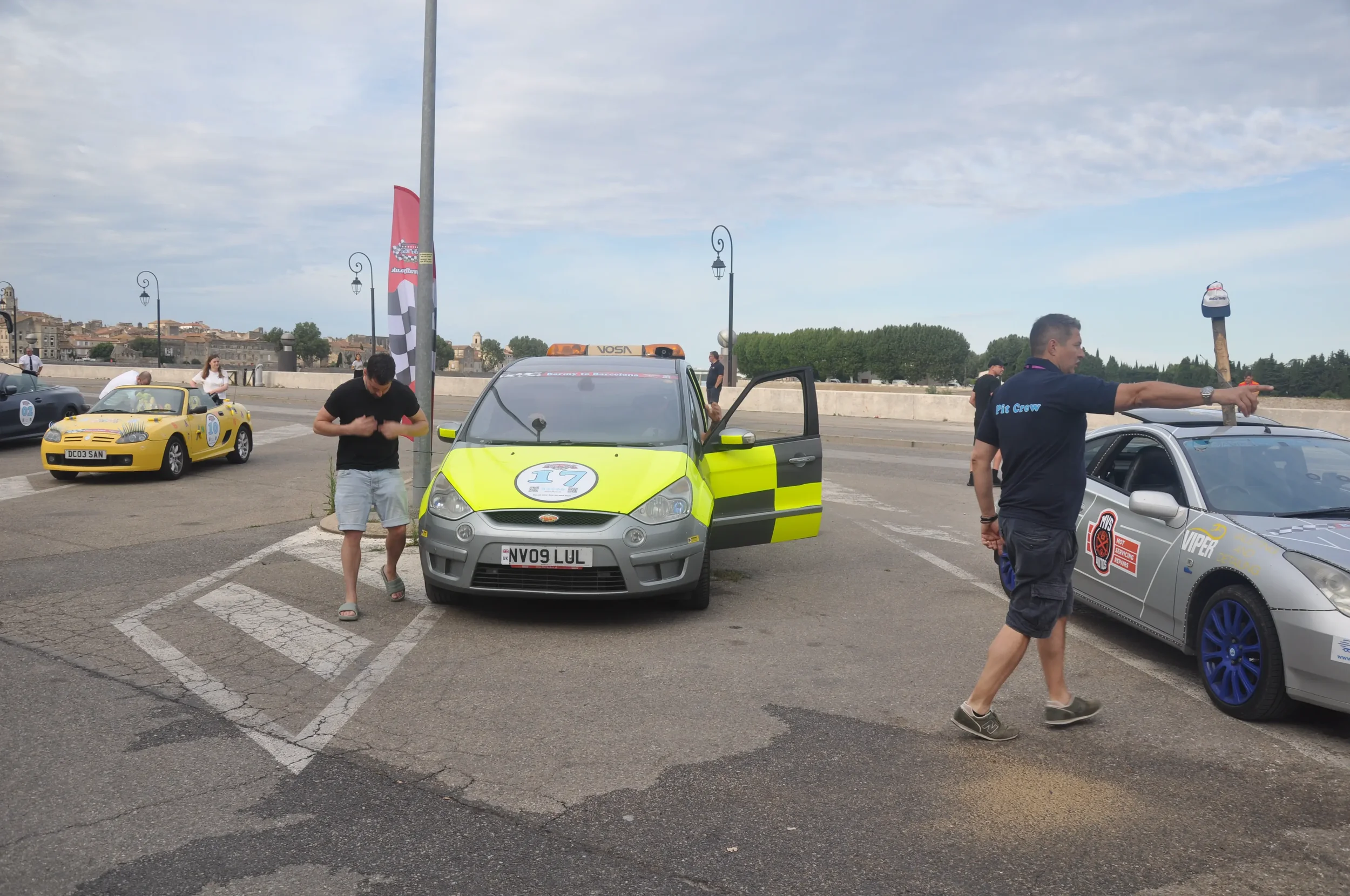 Multiple cars parked in a lot with people around, a yellow sports car, a gray safety car with a license plate NV09 LUL, and a silver Nissan with blue rims; one person is pointing with a stick, and another is preparing to drive.