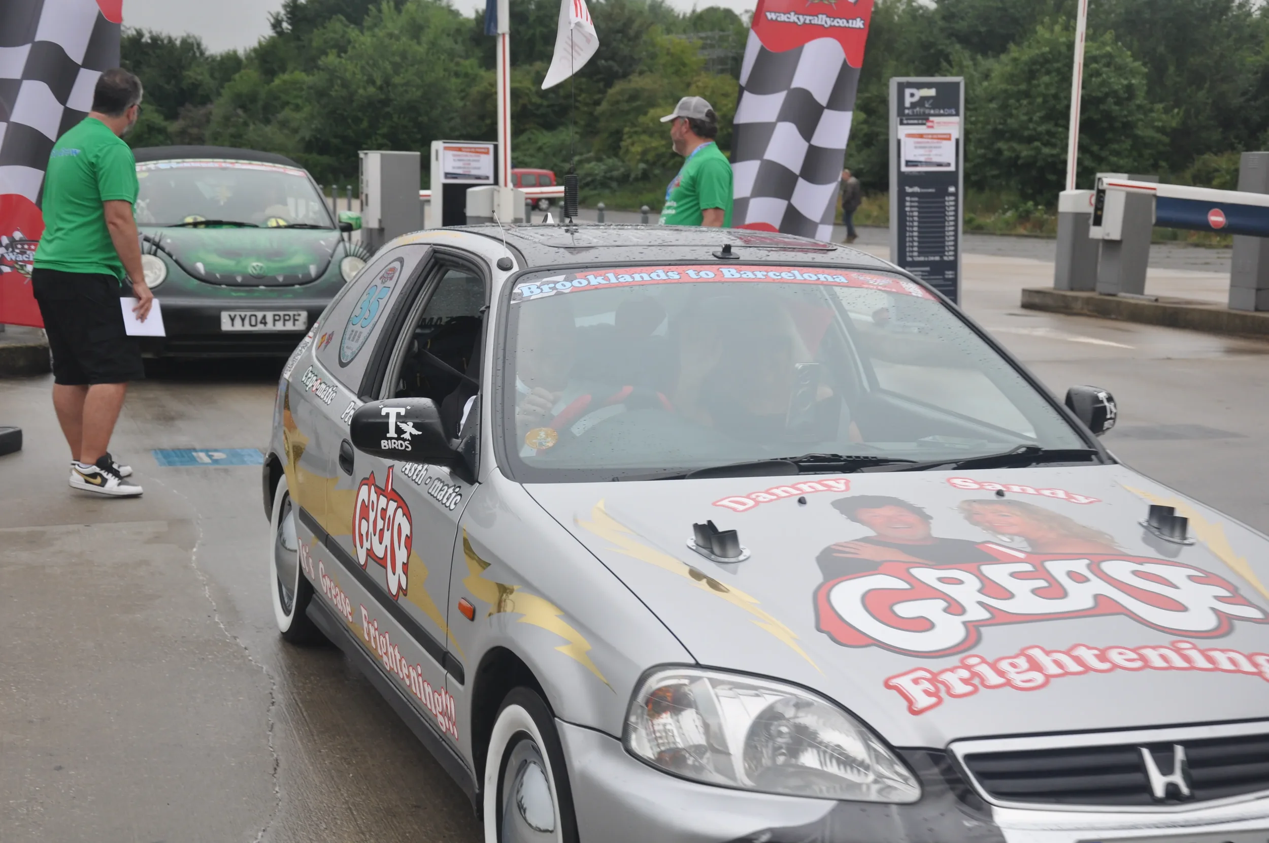 A silver Honda car with advertising decals parked at a rally checkpoint, with three people in green shirts nearby and checkered flags in the background.
