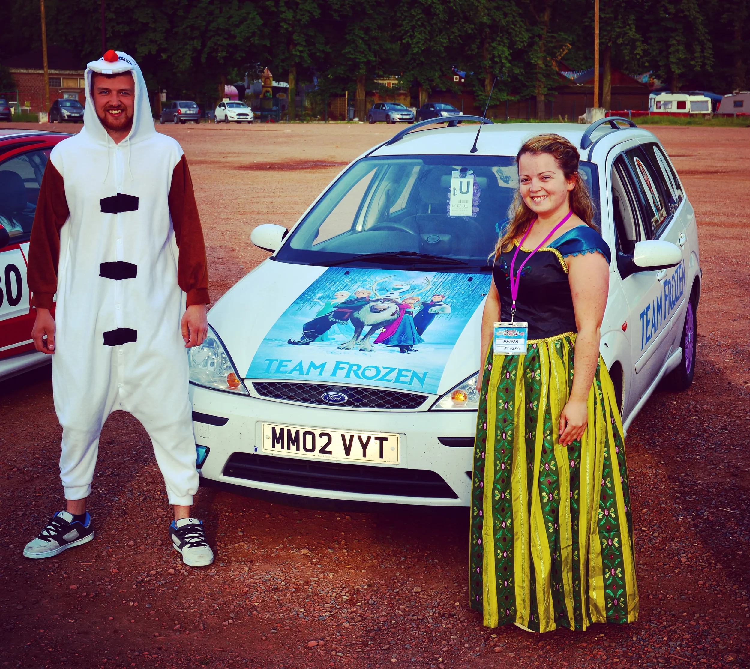 A man in a Olaf from Frozen costume and a woman in a colorful dress standing next to a car with a 'Frozen' themed decal on the hood, at an outdoor event.