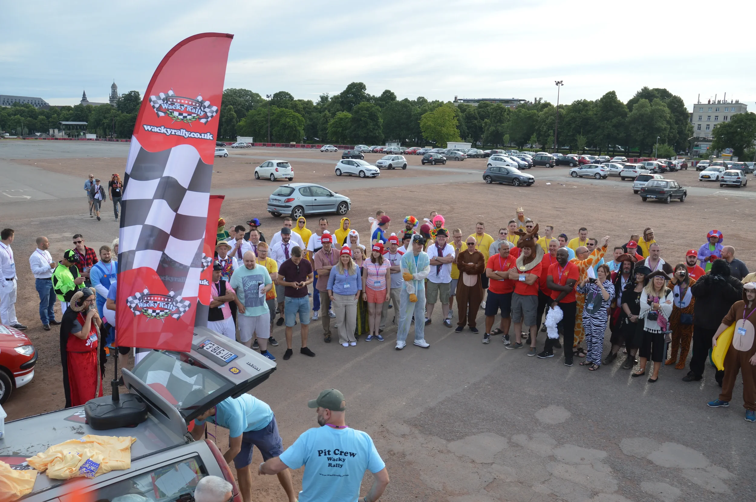 Group of people dressed in colorful costumes and casual clothes gathered outdoors near a Wacky Races event banner with a checkered flag design, in a large open parking area with cars and trees in the background.