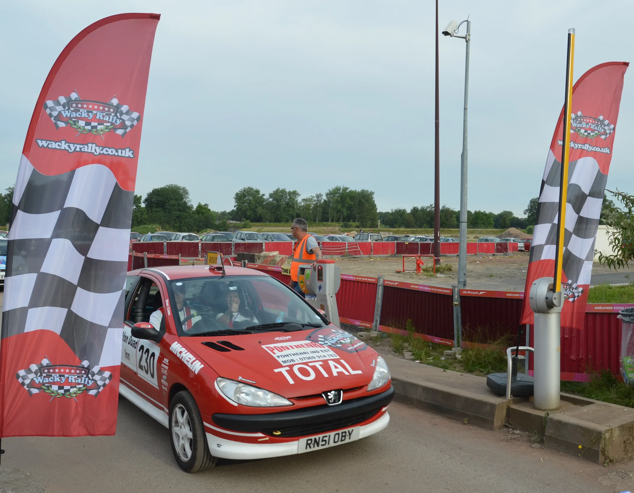 A red rally car with the number 230, sponsored by Total and Michelin, waiting at a checkpoint with flags displaying Wacky Rally logos on either side. A person in a safety vest is near the car, and there are parked cars and a cloudy sky in the backgro