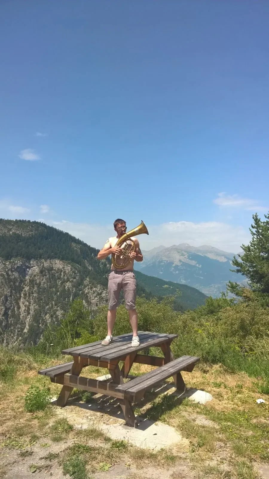 A man standing on a picnic table outdoors playing a saxophone with a mountain landscape in the background under a clear blue sky.