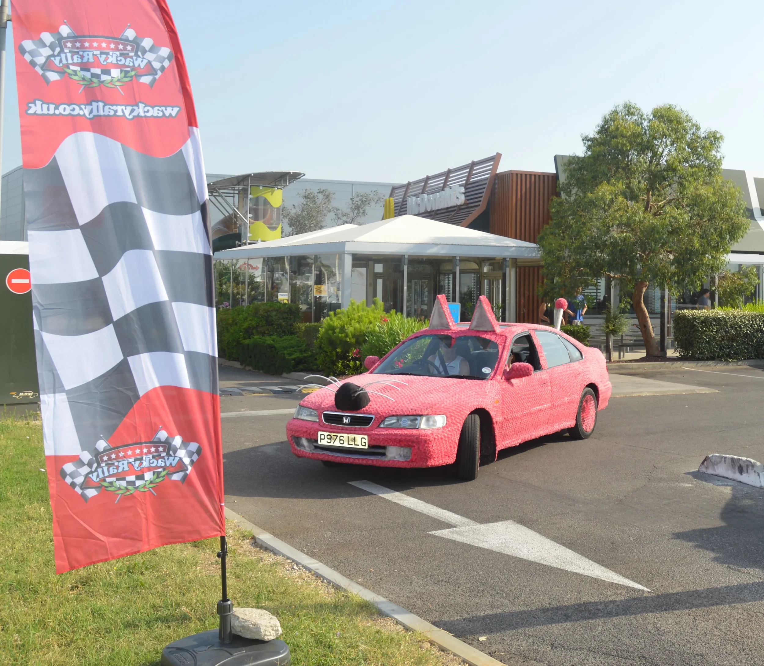 A car decorated to look like a pink mouse with cat ears, a black nose, and whiskers, parked in a lot near a flag with a racing theme and a building in the background.
