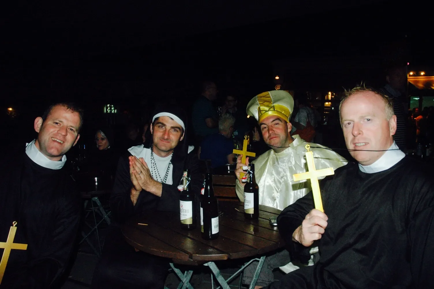 Group of four men dressed in religious and carnival costumes sitting at a table holding crosses and beer bottles, smiling at the camera at night.